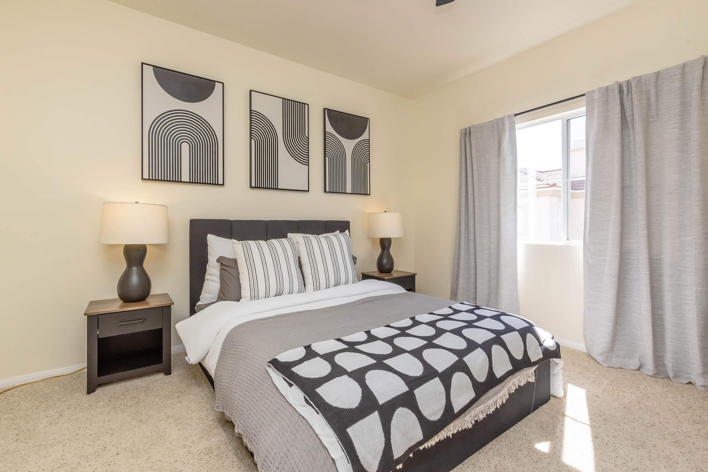 A modern bedroom featuring a queen-sized bed with gray and white bedding, two bedside tables with lamps, and three abstract wall art pieces. Natural light enters through a window with sheer curtains, highlighting the neutral color scheme and carpeted floor.