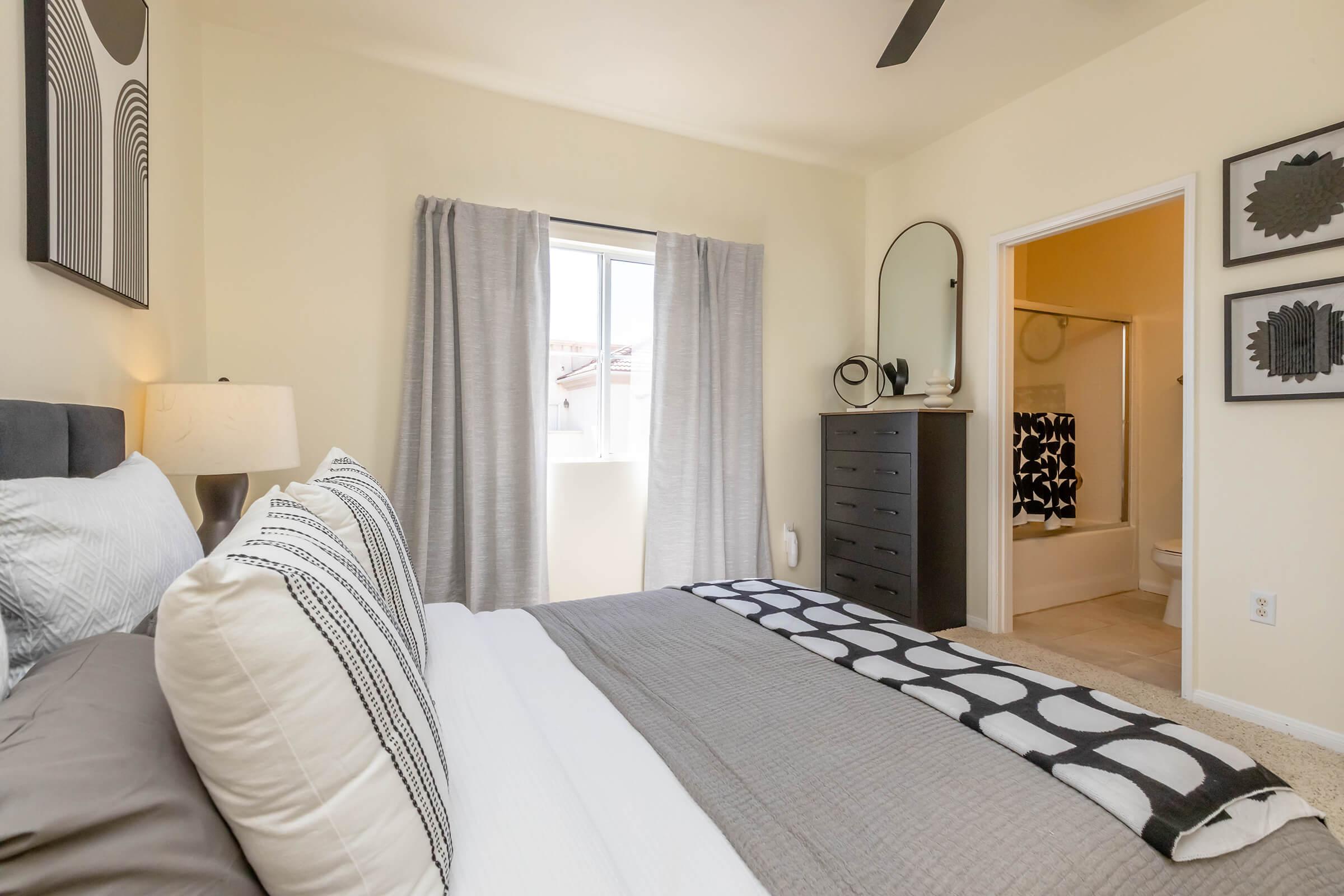 A neat and modern bedroom featuring a gray bedspread with decorative pillows, a bedside lamp, and a dark dresser. Light-colored walls and curtains brighten the space, which has a window revealing a glimpse of the outdoors. A bathroom can be seen through an open door, adding practicality to the room.