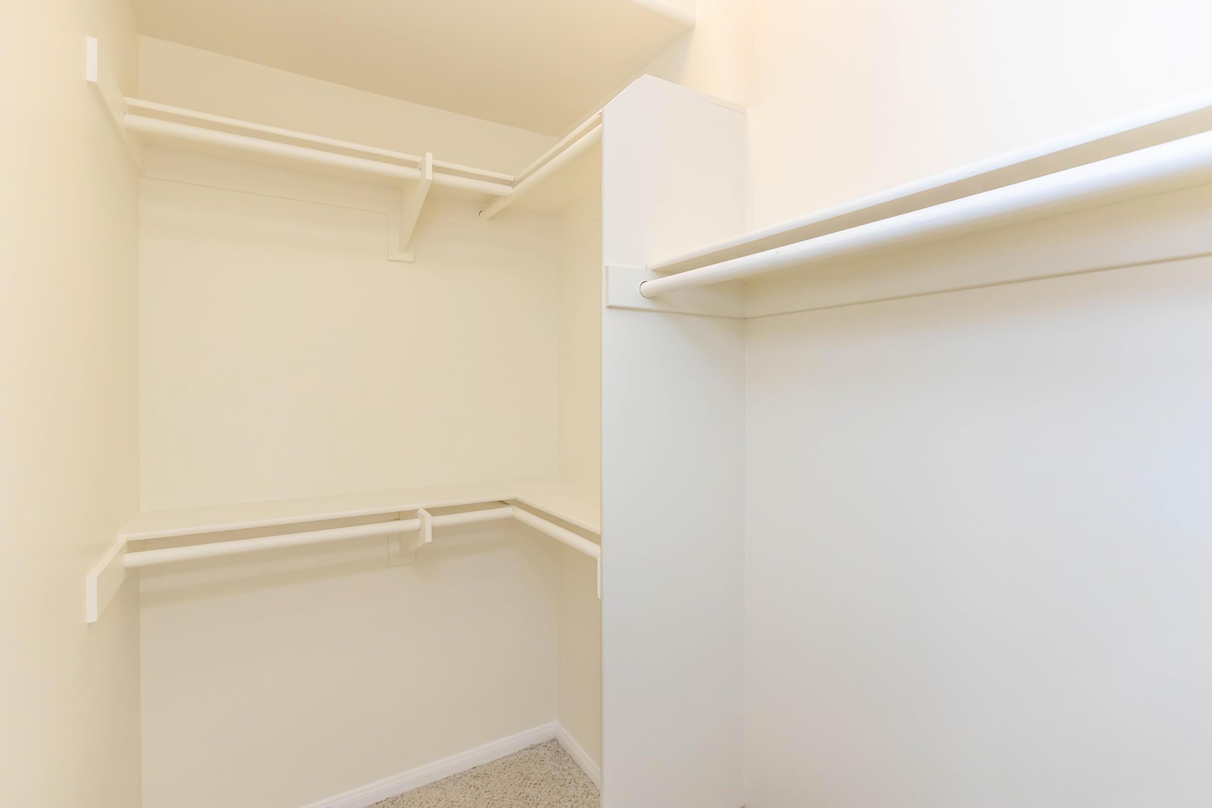 A clean, empty closet with white walls and shelves. The space features two hanging rods and a light beige carpeted floor, creating a minimalist and organized appearance.