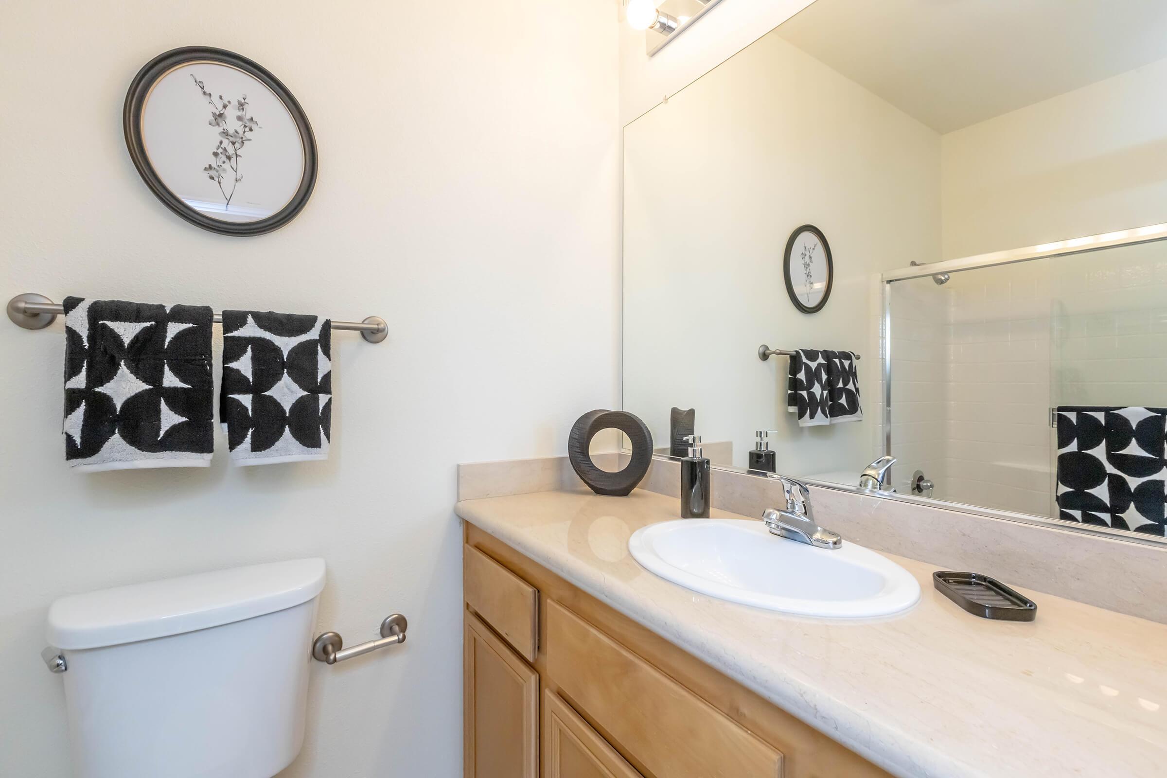 A modern bathroom featuring a sleek white sink with a contemporary faucet, a large mirror, beige cabinetry, and decorative items. Stylish black and white towels hang on a towel rack, complemented by minimalistic decor, including a round wall art piece and a circular container on the countertop.