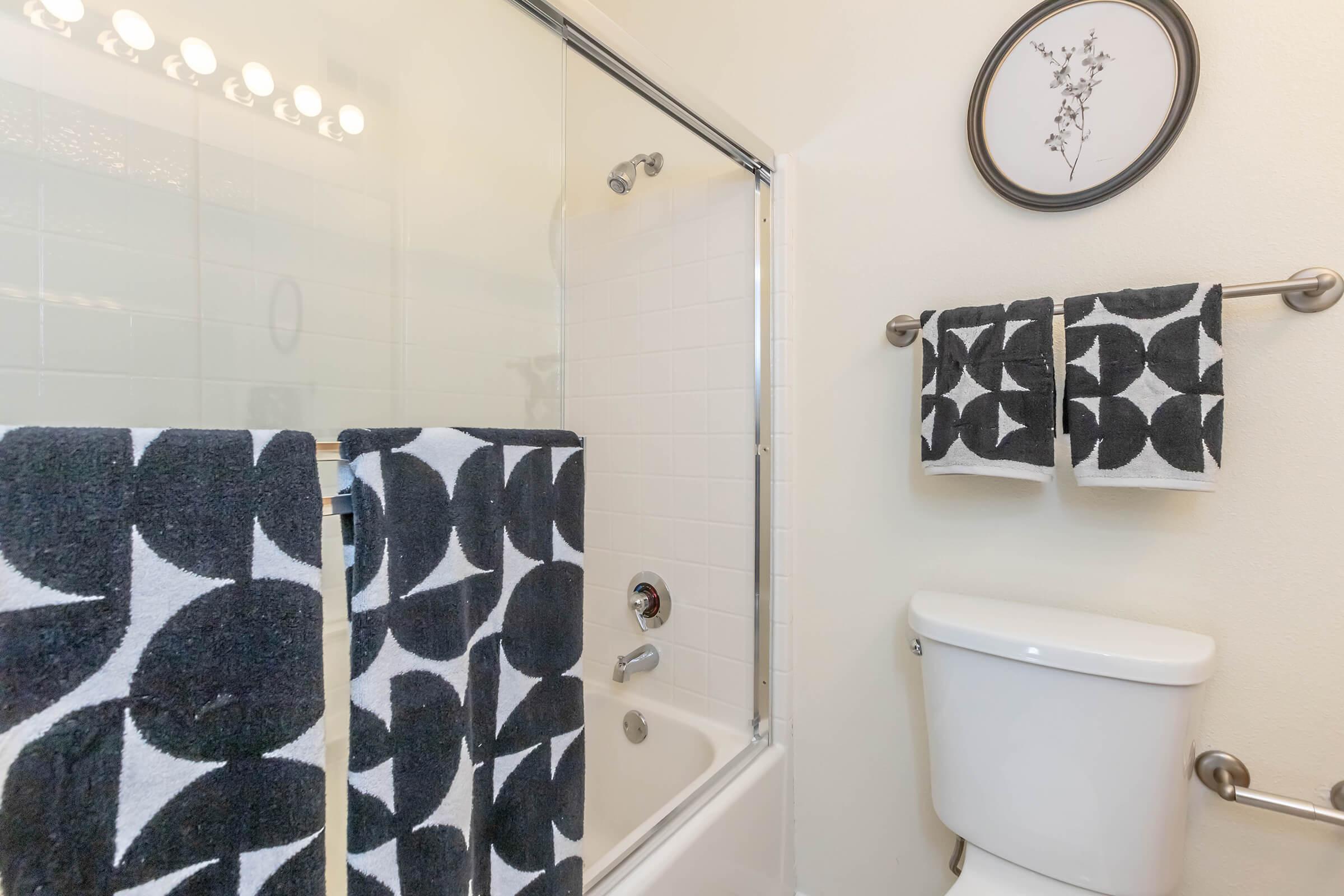 A clean bathroom featuring a shower with glass doors, a white toilet, and black and white patterned towels hanging on a towel rack. A small framed artwork is displayed on the wall. The overall decor is modern and minimalistic, with a light color scheme.