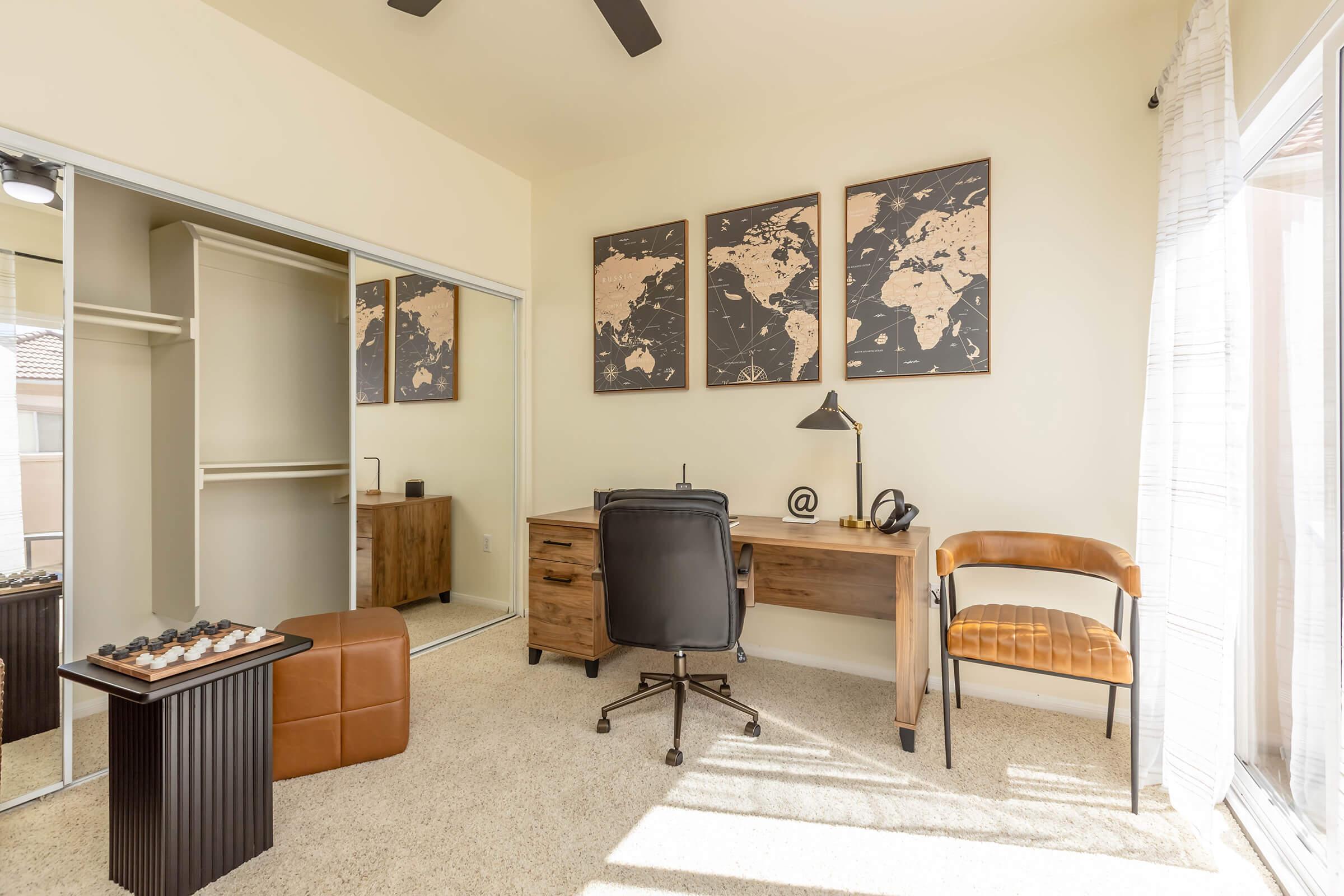 A cozy home office featuring a wooden desk with a black chair, a wall with framed world maps, and a stylish tan chair. The space has a large closet with mirrored doors, a small table with decorative items, and a window letting in natural light, creating a warm and inviting atmosphere for work or study.
