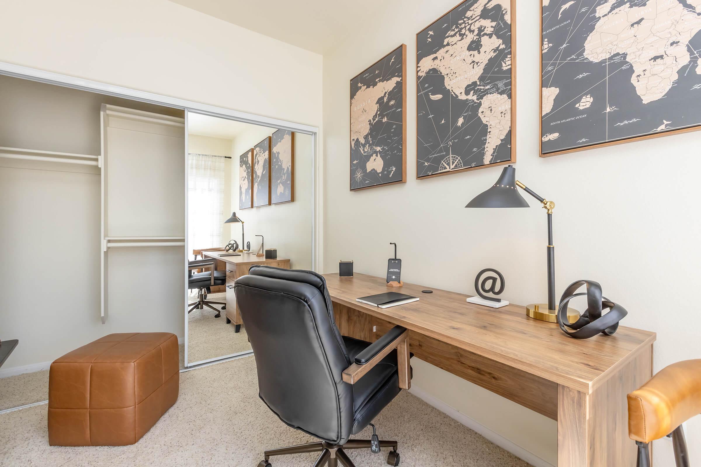 A modern home office setup featuring a wooden desk with a black chair, a desk lamp, and decorative items. Behind the desk are three world map prints hanging on the wall, and a large mirror reflecting a closet space. There's a brown ottoman in the foreground, enhancing the cozy atmosphere.