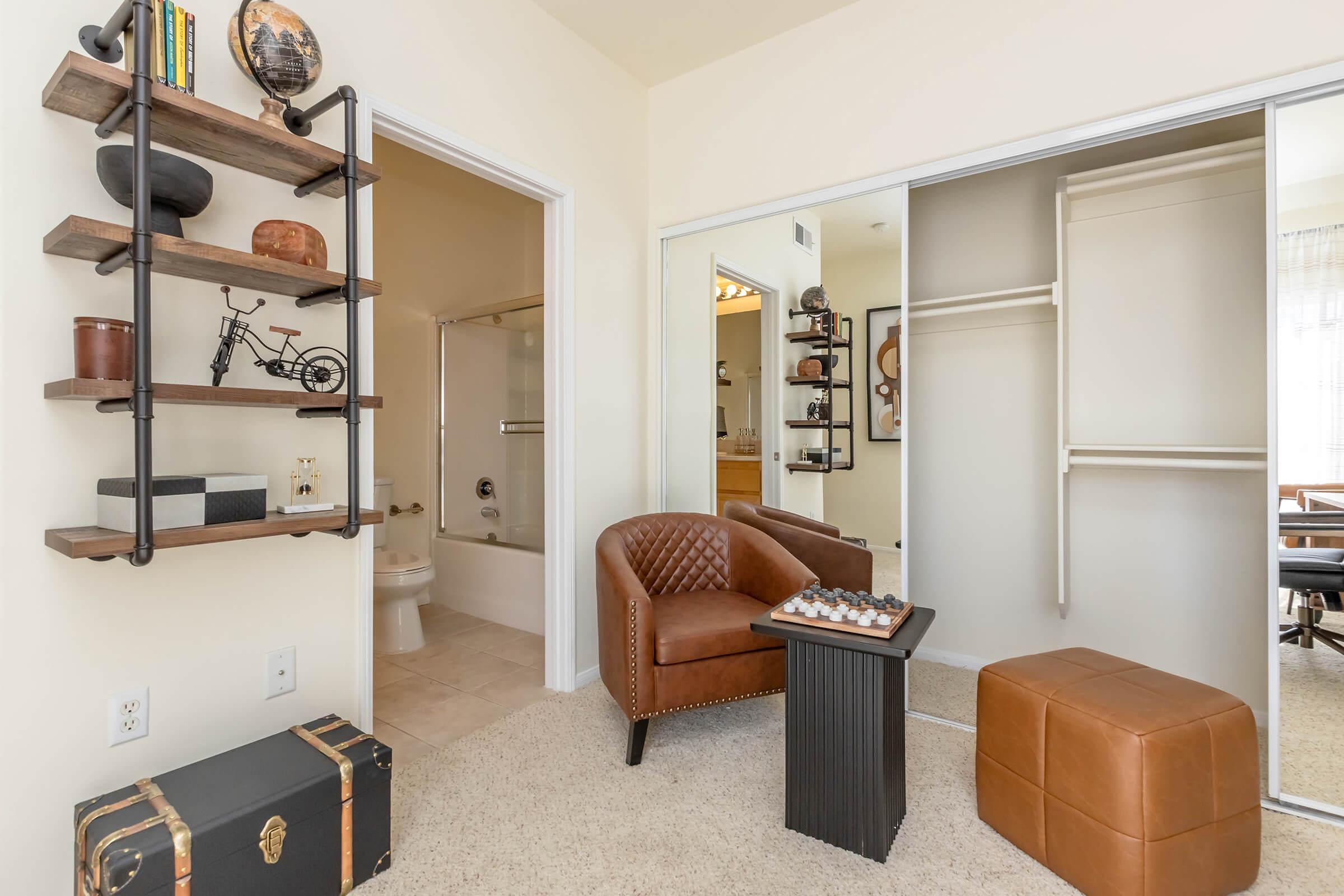 A cozy room featuring a brown leather armchair, a small black table with a decorative item, and a brown ottoman. There’s a stylish shelf on the left with various decor items, and a closet with sliding doors in the background. A bathroom entrance is visible through the doorway.