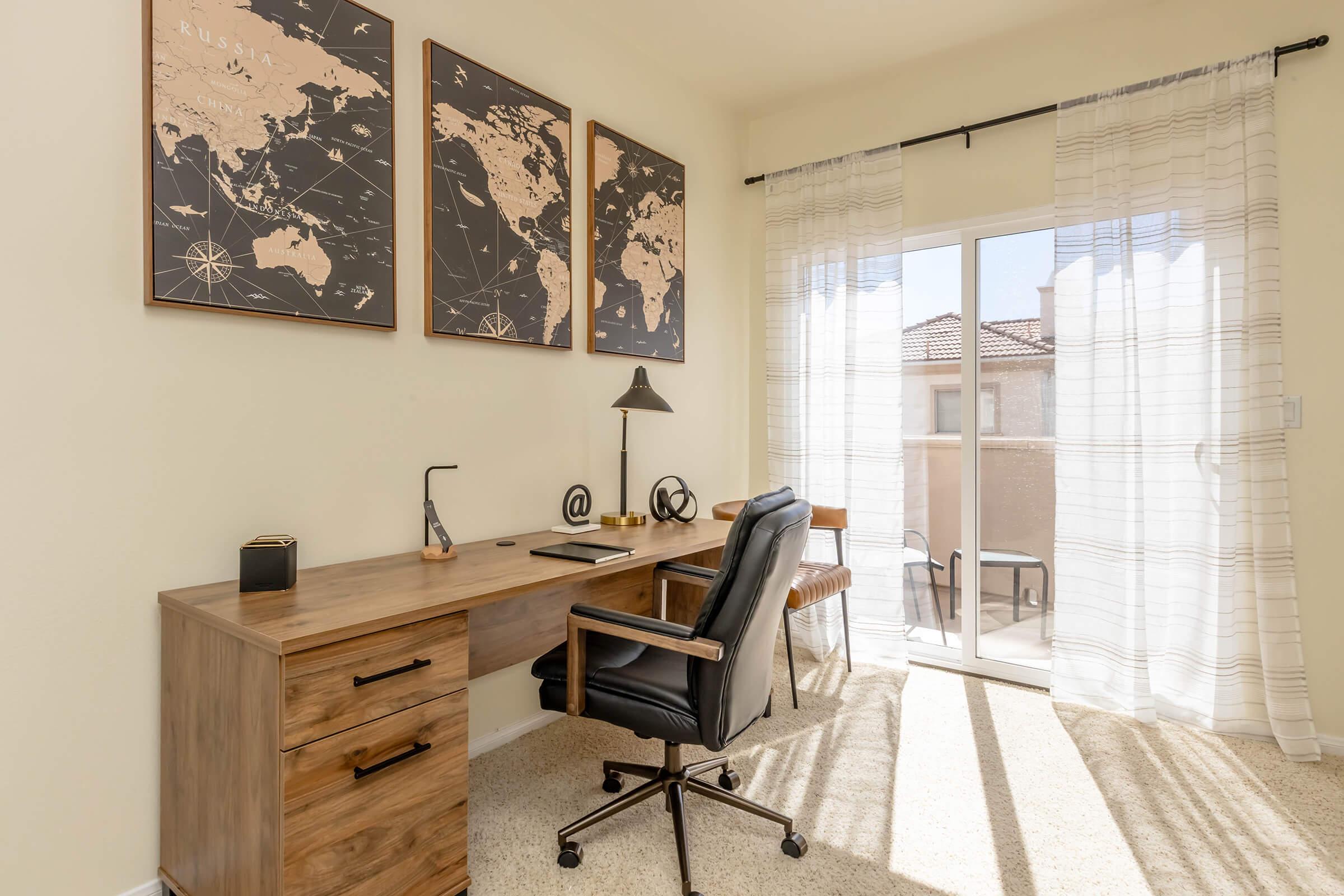 A bright home office featuring a wooden desk with a black ergonomic chair, a modern lamp, and decorative items. The wall displays three framed maps, and large sliding glass doors lead to a balcony, allowing natural light to fill the space. The room has light-colored walls and a soft carpet.
