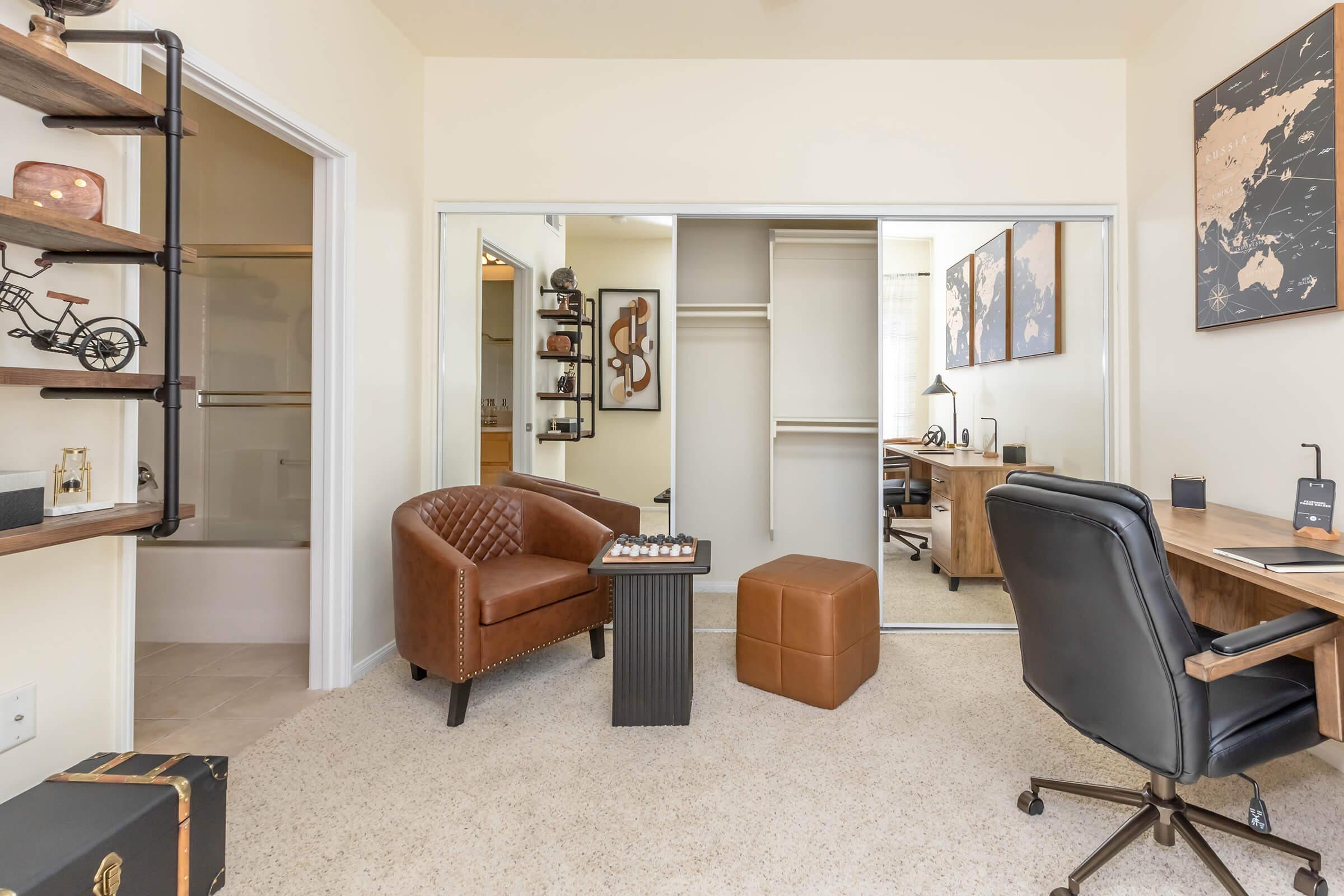A stylish home office featuring a tan leather chair, a small brown ottoman, and a desk with a lamp. The room has a light-colored carpet, and mirrored closet doors reflecting the space. Wall art and decorative shelves add a modern touch to the cozy workspace.