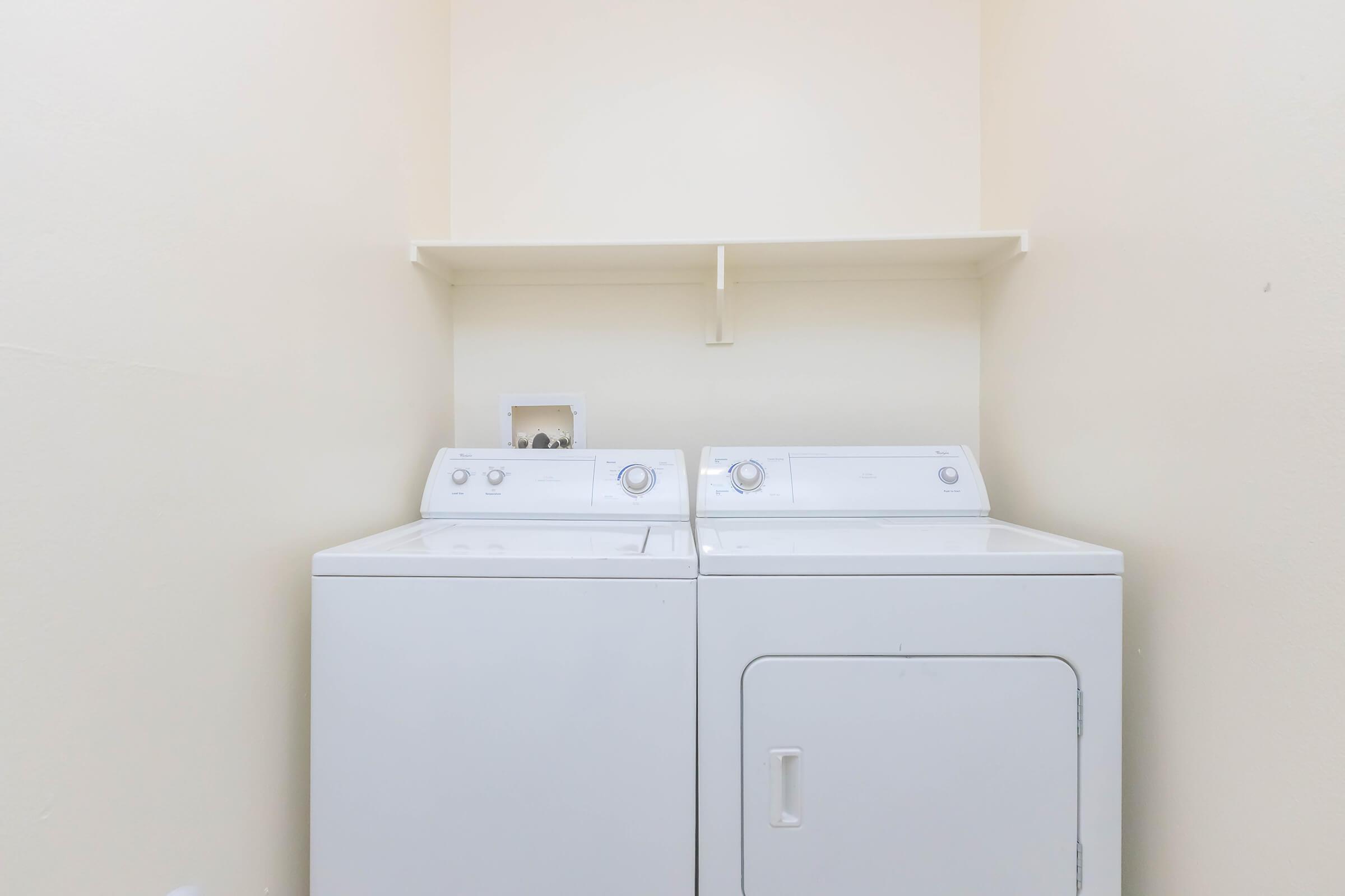 A laundry room featuring a stacked washing machine and dryer. The appliances are white and sit against a plain, light-colored wall. A simple shelf is mounted above the units, providing storage space. The overall atmosphere is clean and functional, suitable for laundry tasks.