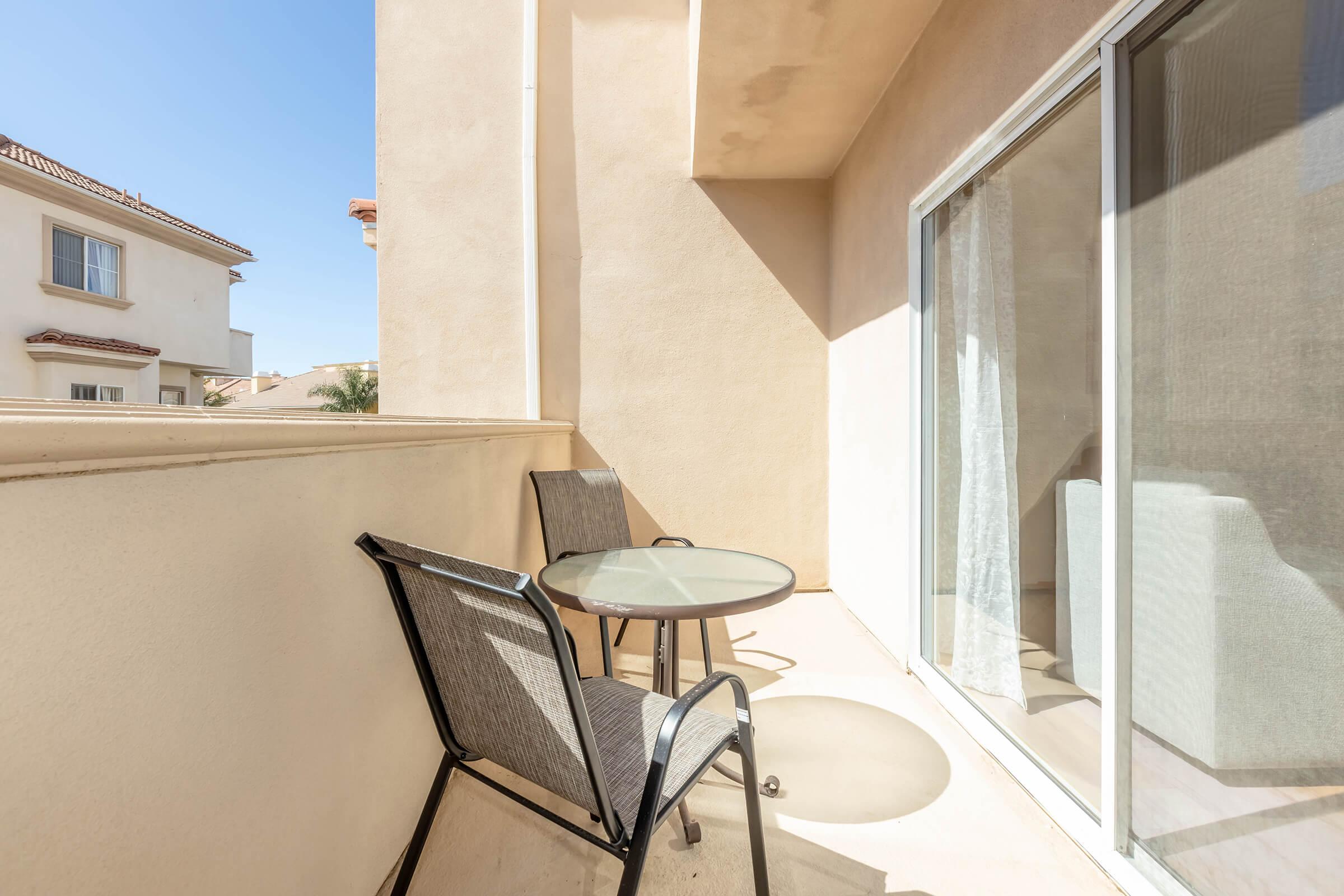 A small balcony featuring two metal chairs and a round glass table, with sunlight casting shadows on the floor. The view includes neighboring buildings and a clear blue sky.