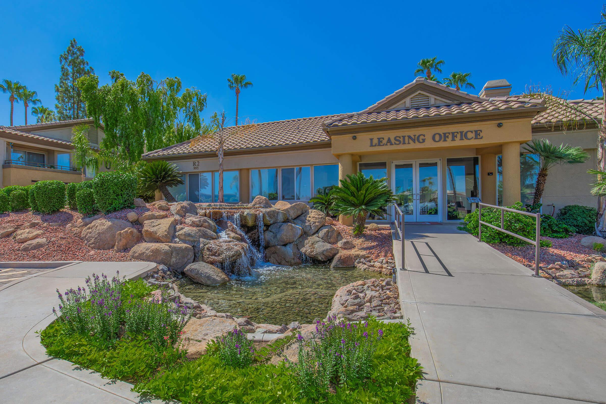 A leasing office building surrounded by lush landscaping, including palm trees and colorful flowers. A rock formation with flowing water is featured in front of the office, with a clear blue sky overhead, creating a welcoming and serene atmosphere.