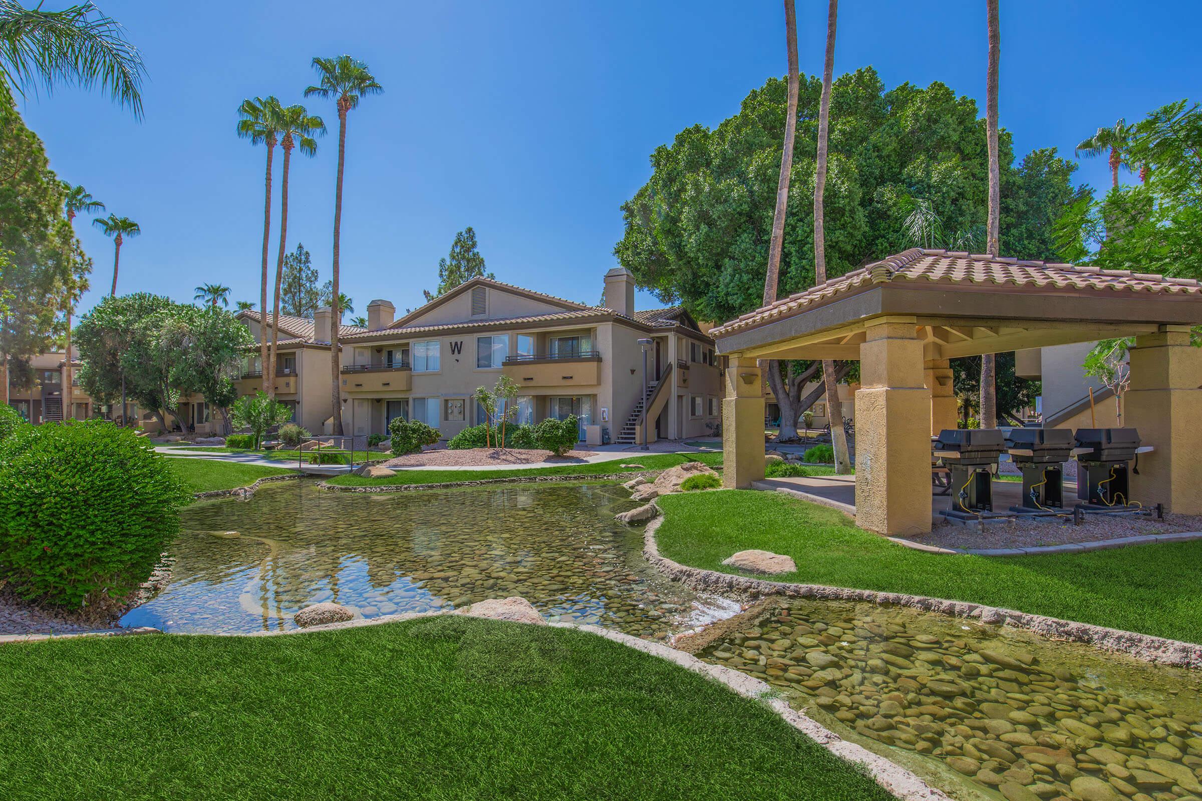 A serene residential complex featuring two-story buildings with a fountain and a small pond surrounded by lush green lawns. Palm trees and shrubs add to the tropical ambiance, while a shaded area with grills invites outdoor gatherings. Bright blue sky enhances the peaceful atmosphere.