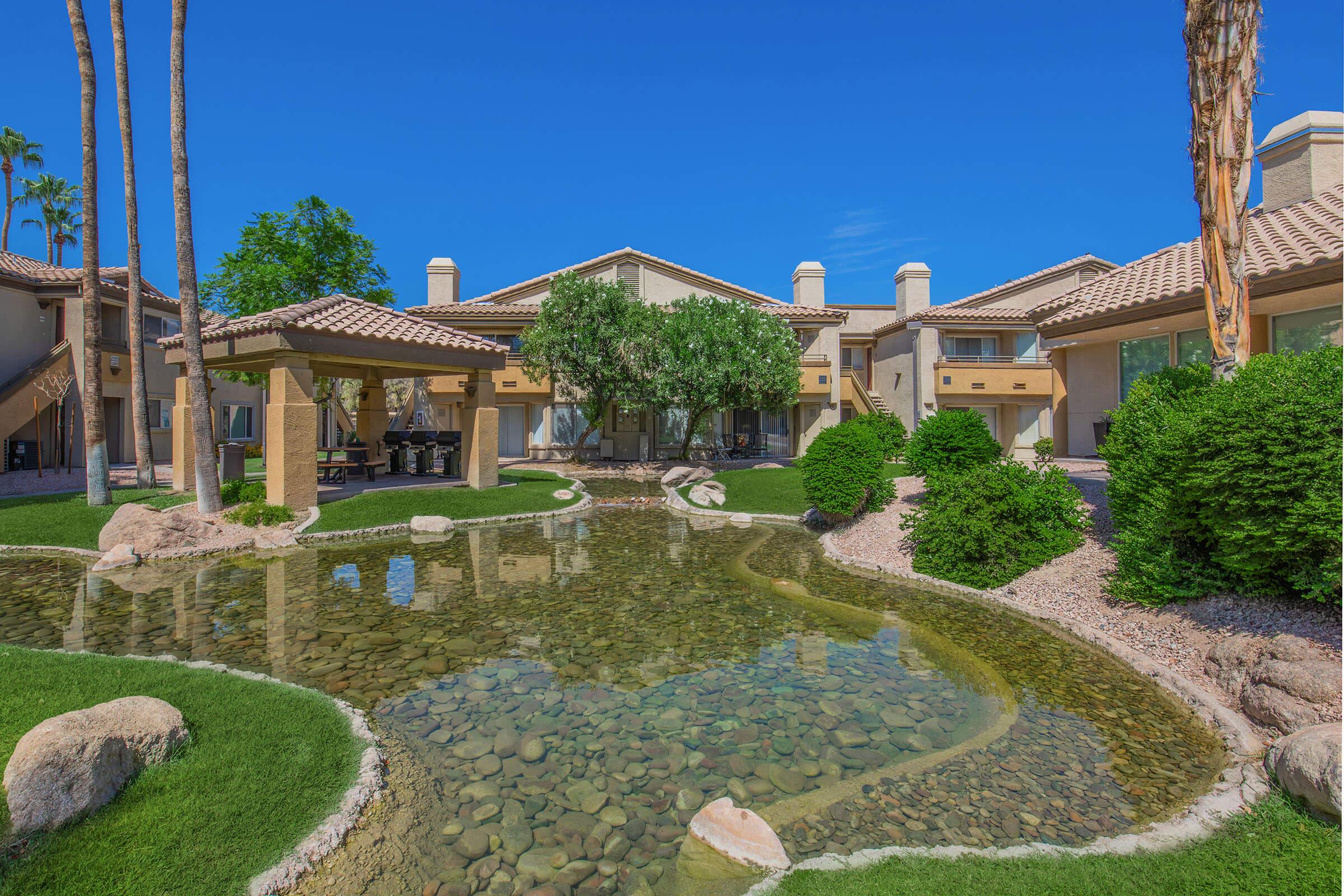 A serene courtyard with a winding pond surrounded by lush greenery and decorative rocks. Palm trees stand tall against a blue sky, and nearby buildings feature Mediterranean-style architecture. The scene evokes a peaceful and relaxing atmosphere, perfect for outdoor leisure.