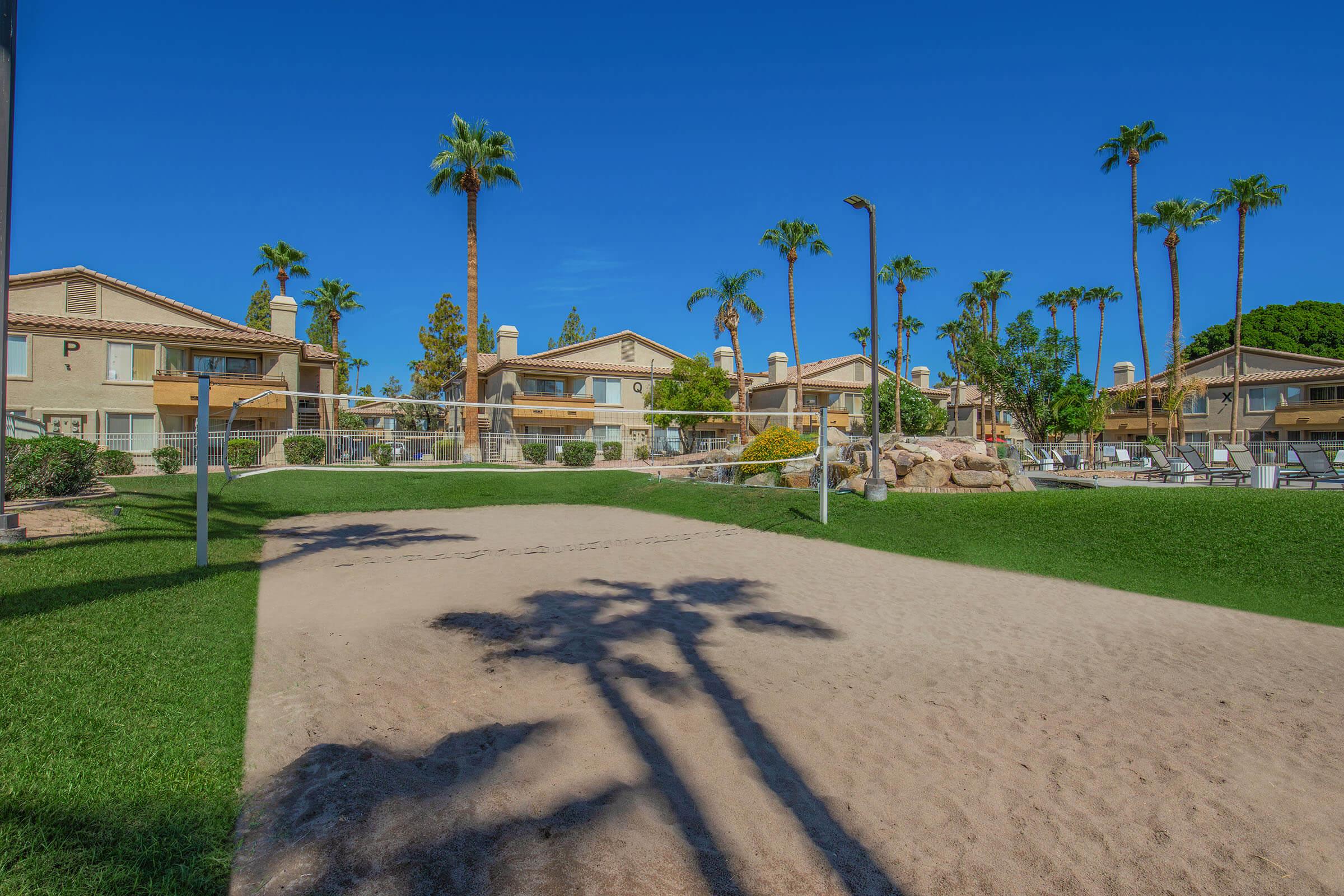 Beach volleyball court with sandy area in foreground, surrounded by lush green grass. In the background, there are several multi-story residential buildings and tall palm trees under a clear blue sky. Sun is shining, creating a vibrant and inviting atmosphere.