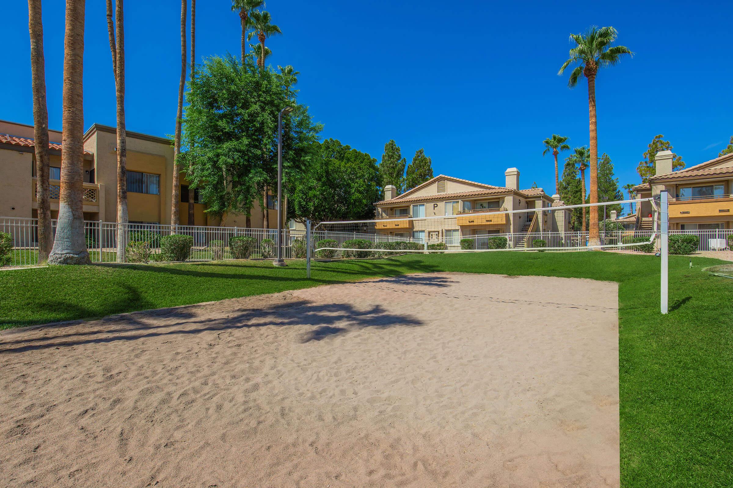 A sandy volleyball court surrounded by lush green grass and palm trees, set against a backdrop of apartment buildings. The clear blue sky complements the serene outdoor setting, ideal for recreation and relaxation.