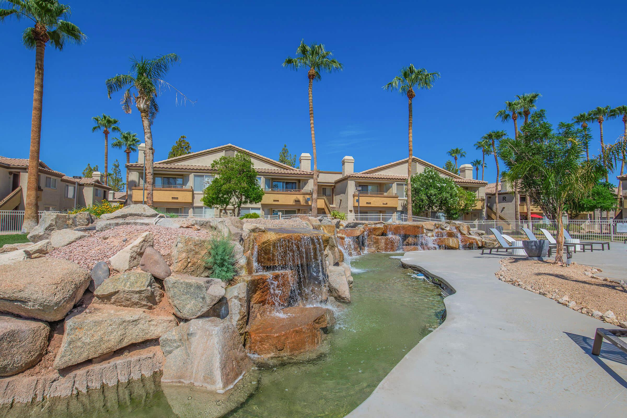 A landscaped area featuring a small waterfall surrounded by rocks, with palm trees in the background. There are several residential buildings with balconies, and a paved pathway leading through the scenery. The sky is clear and blue, enhancing the tropical atmosphere.