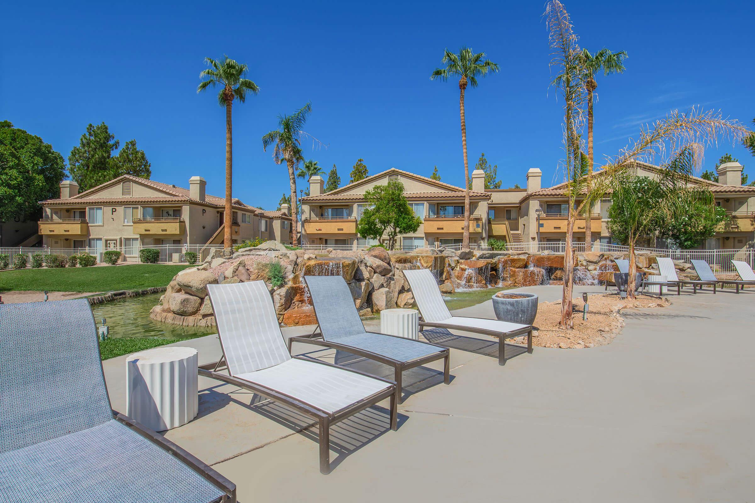 A tranquil pool area featuring empty lounge chairs aligned by a small body of water. In the background, there are palm trees and residential buildings with a Mediterranean style. The sky is clear and blue, creating a relaxing atmosphere.