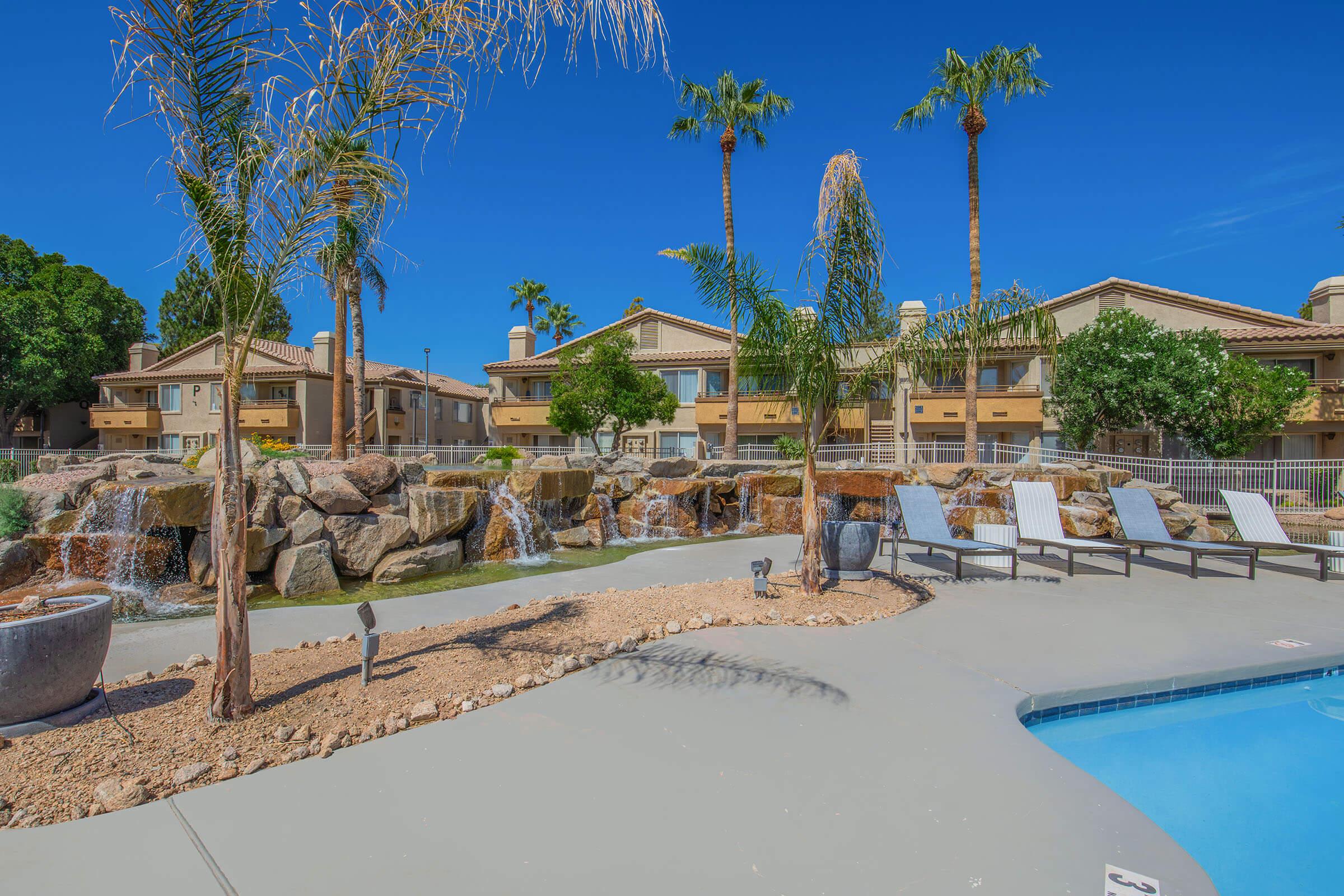 A sunny outdoor pool area featuring lounge chairs, a rock waterfall, and palm trees. In the background, there are residential buildings with balconies surrounded by greenery. The clear blue sky creates a bright, inviting atmosphere.