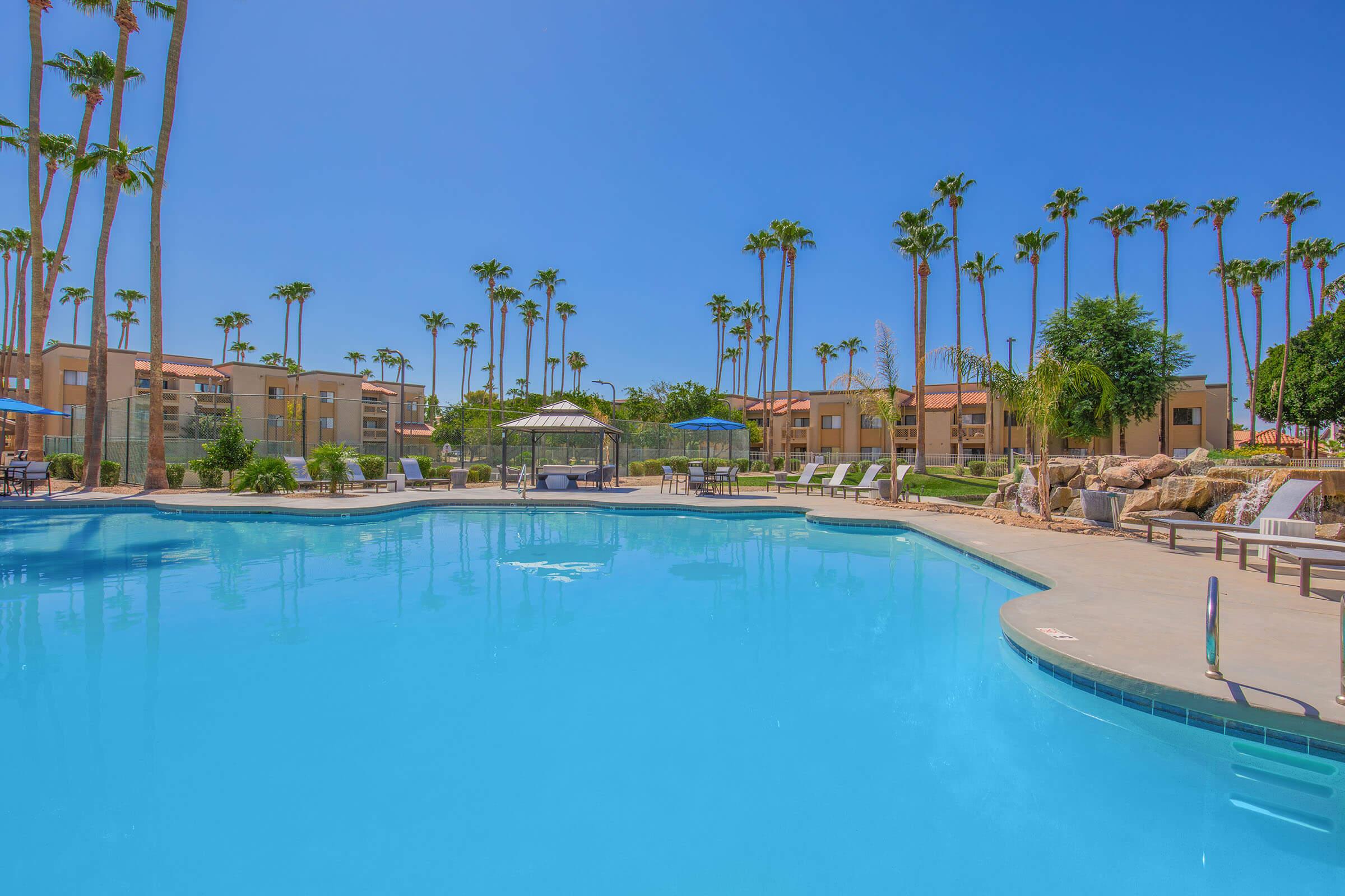 A bright, sunny pool area surrounded by palm trees. The clear blue water reflects the sky, with lounge chairs and umbrellas positioned around the pool. In the background, there are residential buildings and landscaped greenery, creating a relaxing outdoor atmosphere.