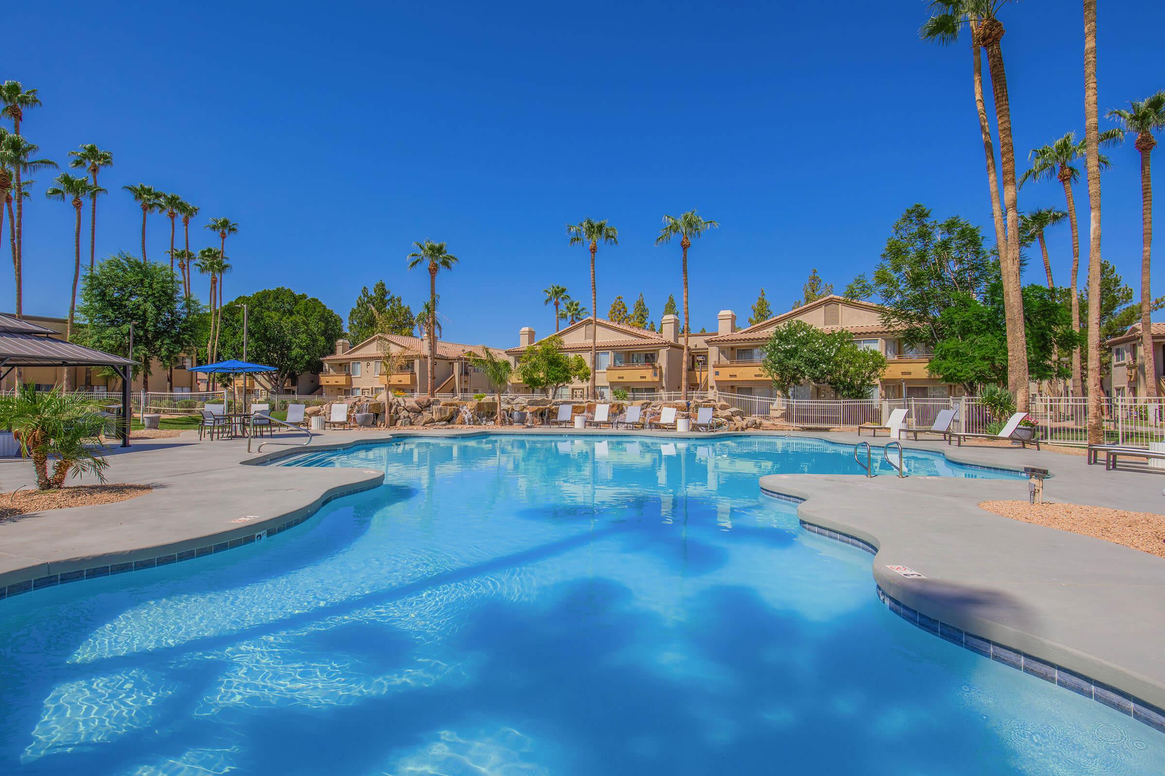 A serene swimming pool surrounded by palm trees and lounge chairs, with residential buildings in the background. The clear blue water reflects the bright blue sky, creating a tranquil atmosphere perfect for relaxation and leisure.