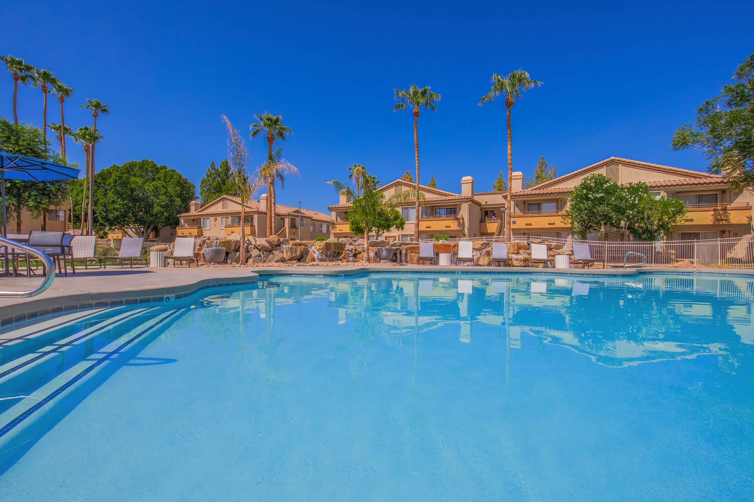 A sparkling blue swimming pool surrounded by palm trees and shaded seating areas. In the background, there are multi-story residential buildings with a sunny sky overhead. The scene conveys a relaxing and inviting atmosphere typical of a resort or leisure community.