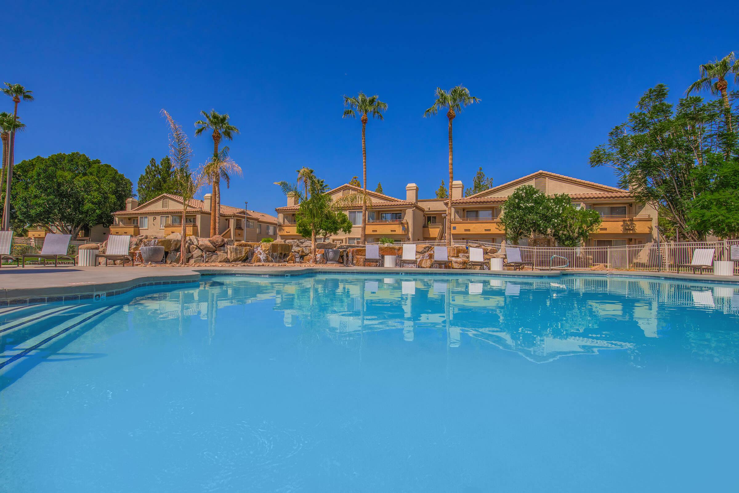 A sparkling blue swimming pool surrounded by lounge chairs, with palm trees swaying in the background. In the distance, residential buildings with a warm color palette are visible under a clear blue sky. The scene conveys a relaxing, sunny atmosphere perfect for a vacation.