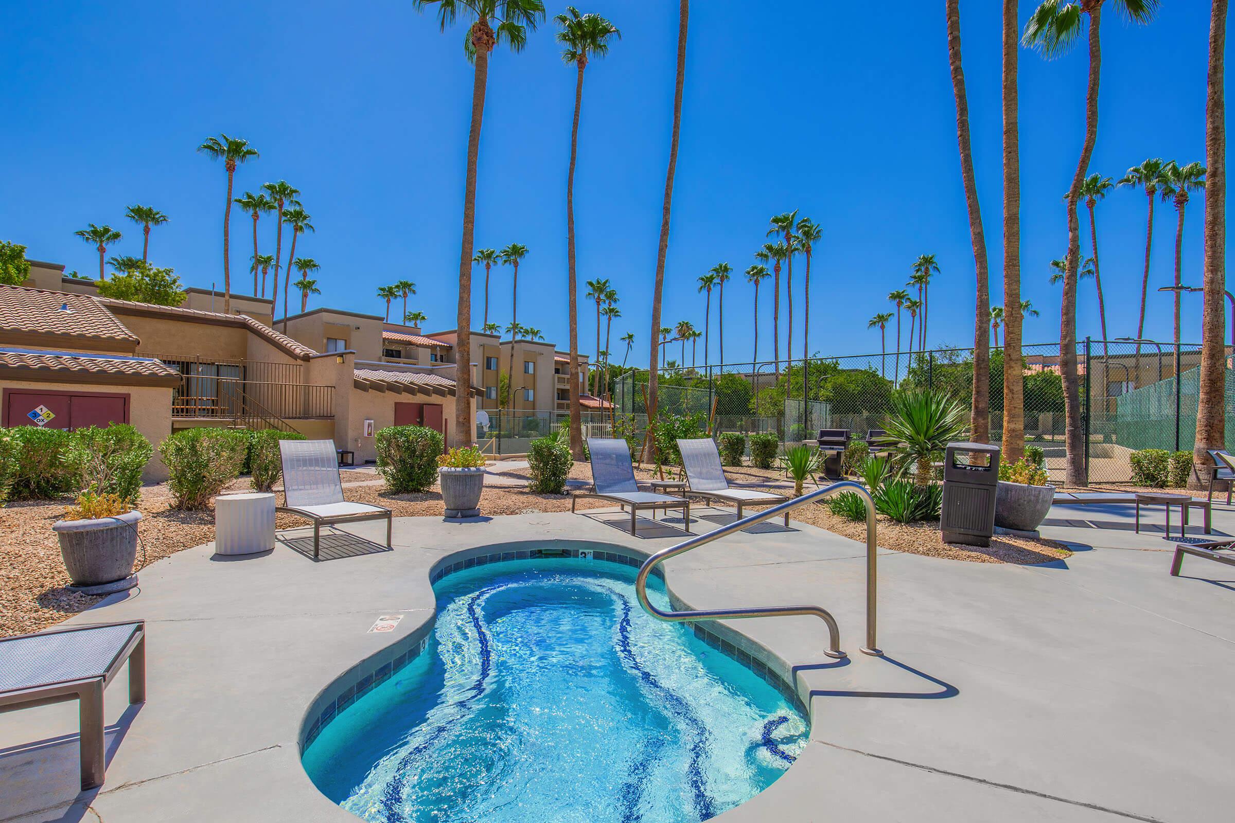 A sunlit pool area featuring a jacuzzi surrounded by lounge chairs, palm trees, and desert landscaping. The clear blue sky adds to the relaxing atmosphere, with nearby buildings in a warm color palette creating a serene and inviting scene.