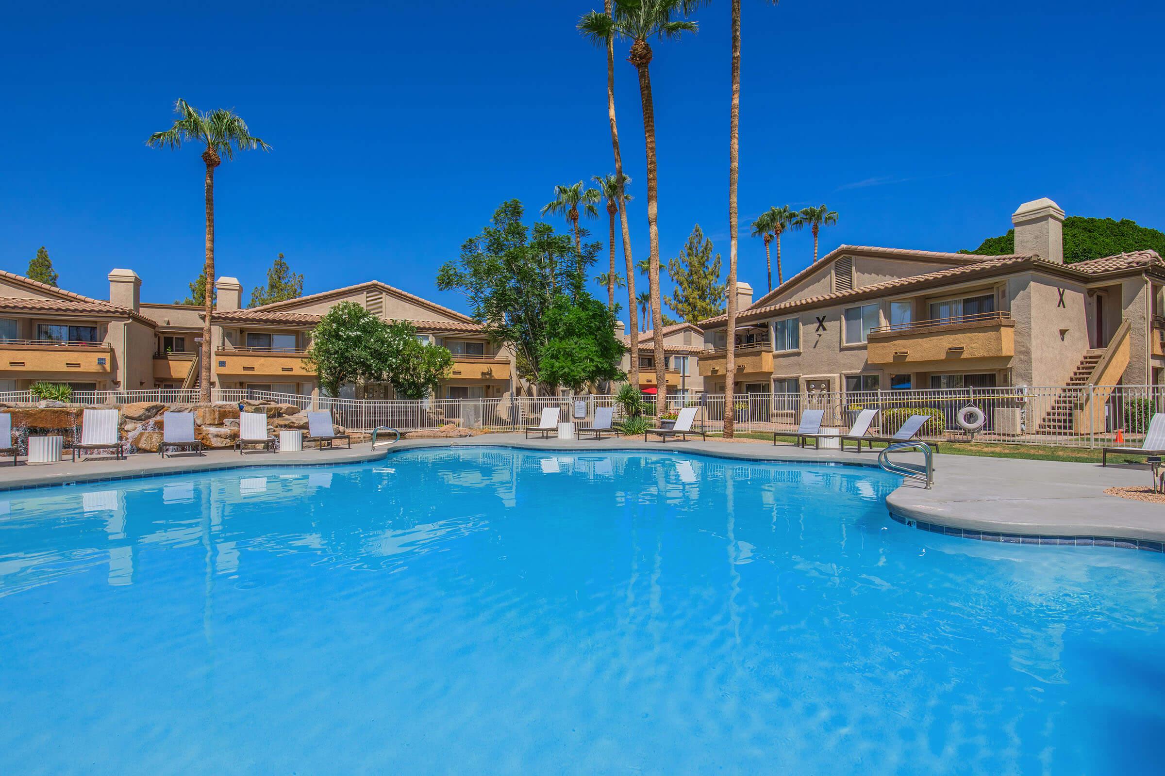 A tranquil swimming pool surrounded by lounge chairs and tall palm trees, with a backdrop of brown multi-story buildings under a clear blue sky. The setting conveys a relaxing vacation atmosphere, ideal for leisure and enjoyment.