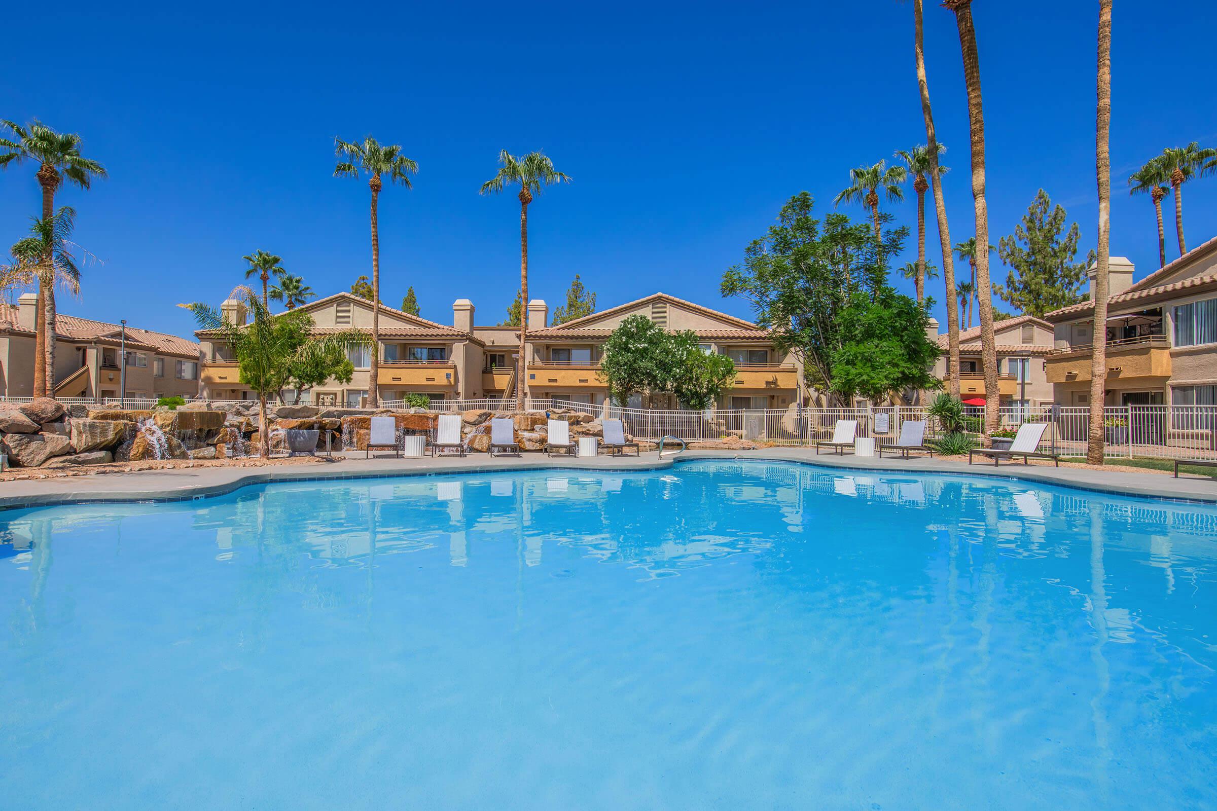 A clear, blue swimming pool surrounded by palm trees and lounge chairs, with residential buildings in the background against a bright blue sky. The scene conveys a relaxed and inviting atmosphere typical of a vacation destination.