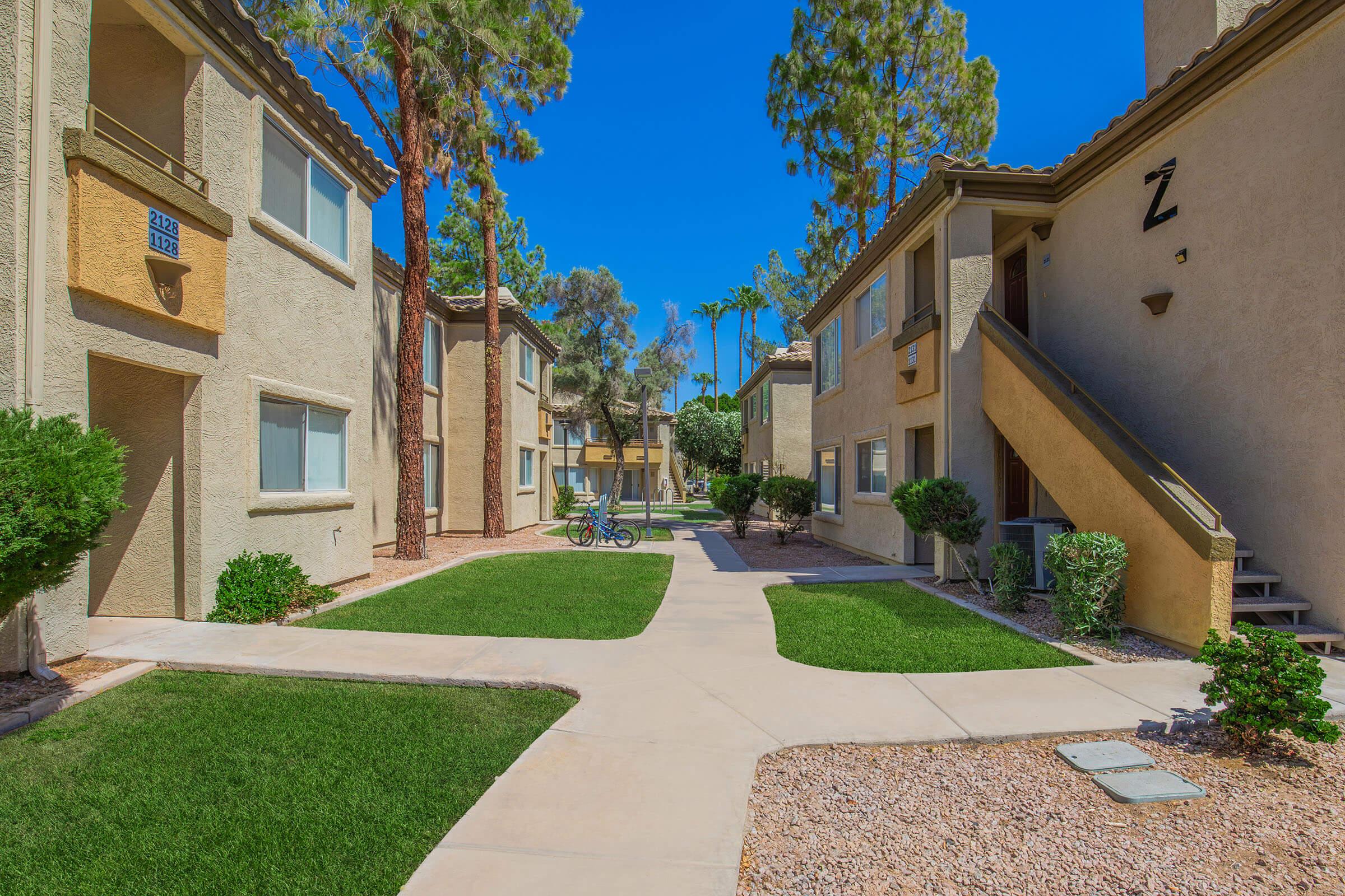 A sunny view of an apartment complex featuring two-story buildings with a landscaped courtyard. Green grass and shrubs line the pathways, and tall trees provide shade. A bicycle is parked in the courtyard, creating a welcoming atmosphere in a residential neighborhood.