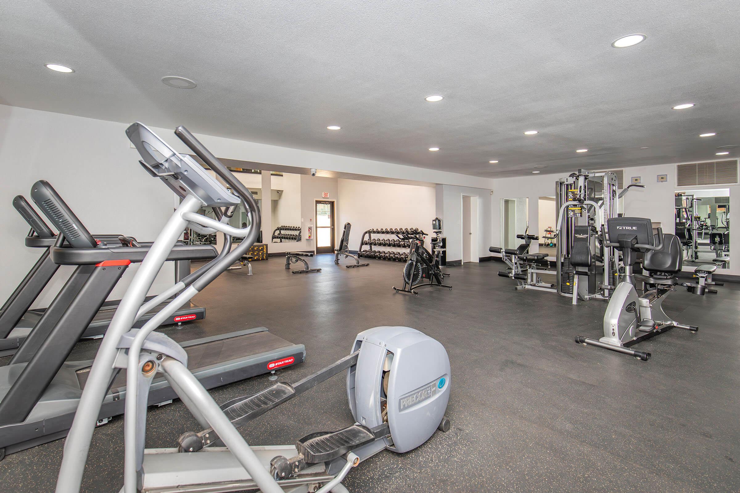 A spacious gym interior featuring various exercise equipment including treadmills, ellipticals, stationary bikes, and weightlifting benches. The floor is dark and the walls are light-colored, creating a bright and open atmosphere for fitness activities.
