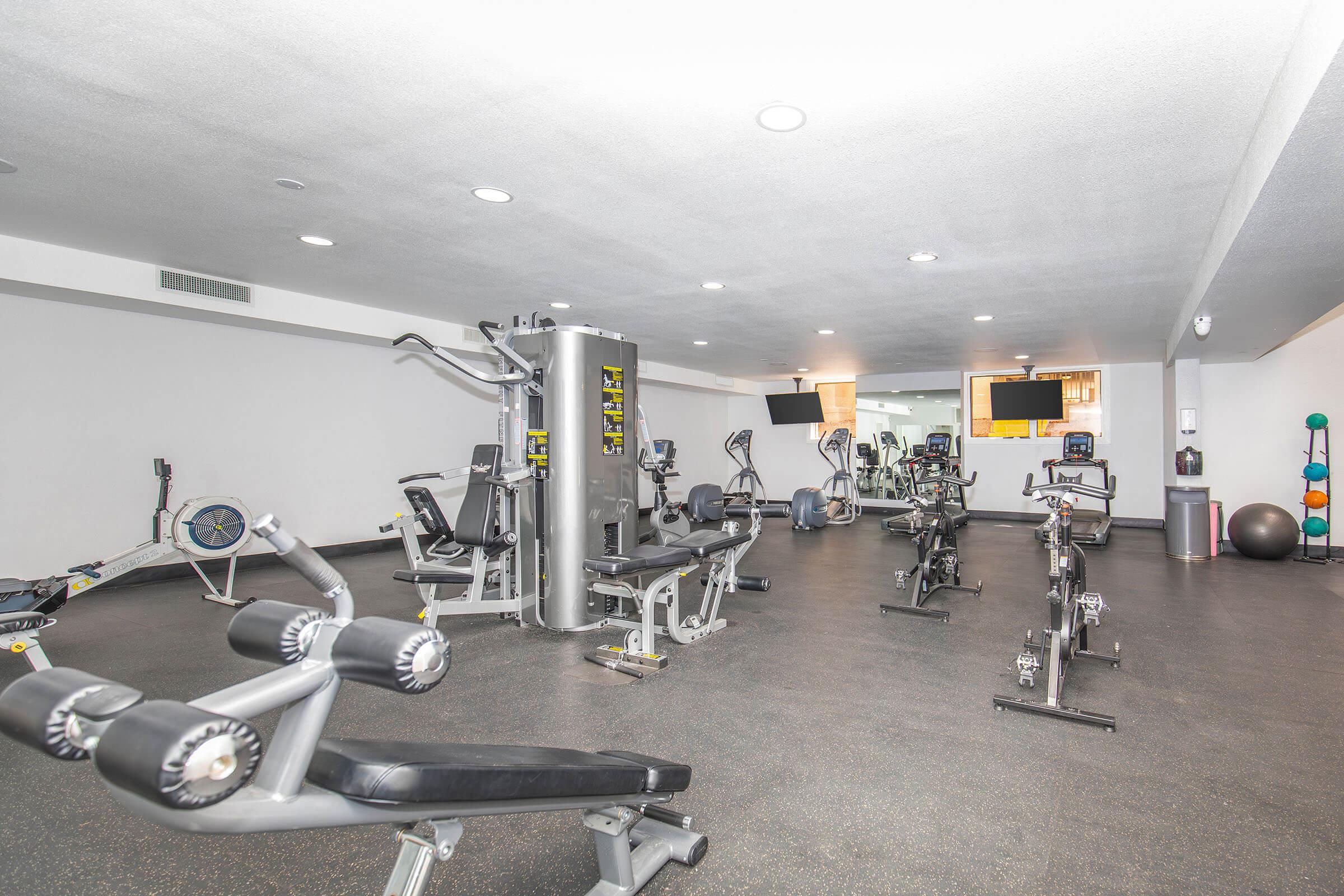 A modern gym with various exercise equipment, including a weight machine, stationary bikes, an elliptical machine, and free weights. The space is well-lit, featuring a minimalist design with smooth flooring and a few mirrors on the walls. Several workout stations are arranged for fitness activities.
