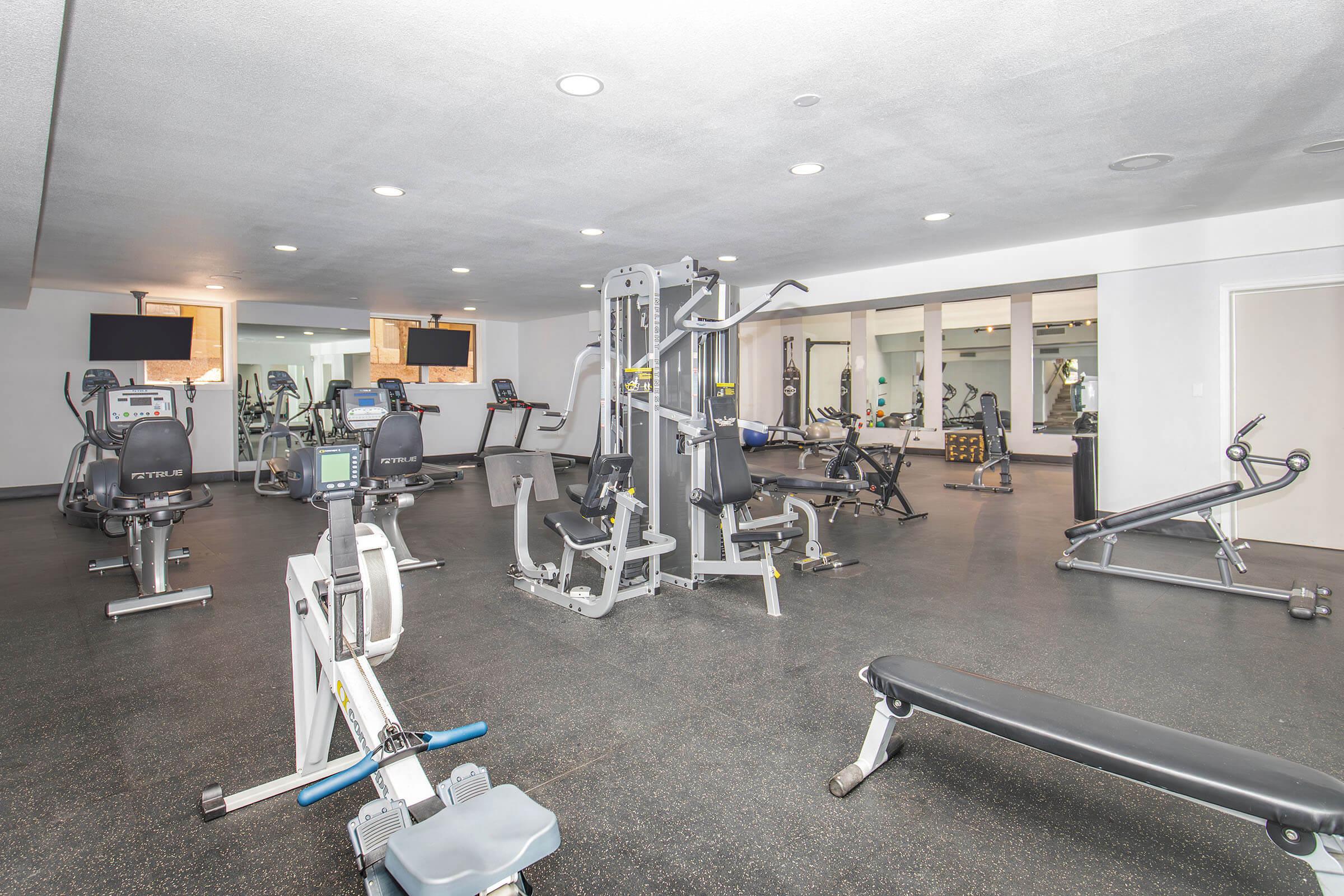 A modern gym interior featuring a variety of workout equipment including treadmills, stationary bikes, weight machines, and benches. The space is well-lit with mirrors on one wall, and there are two TVs mounted on the ceiling. The flooring is dark, and the overall atmosphere is clean and inviting for fitness activities.