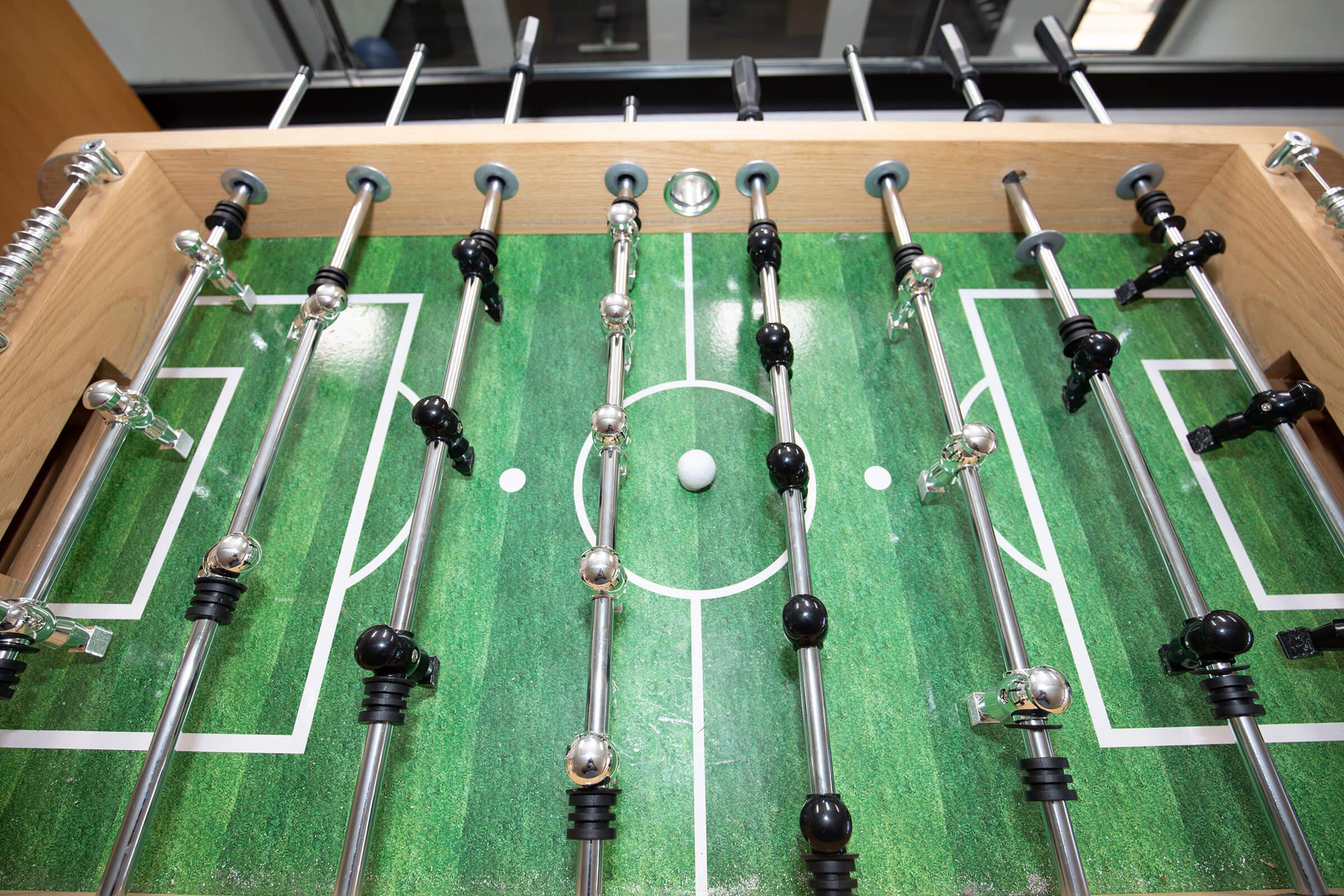 A foosball table with black and silver players positioned along metal rods, set on a green grass-like surface. A small white ball is centered on the field, with the table surrounded by a wooden frame.