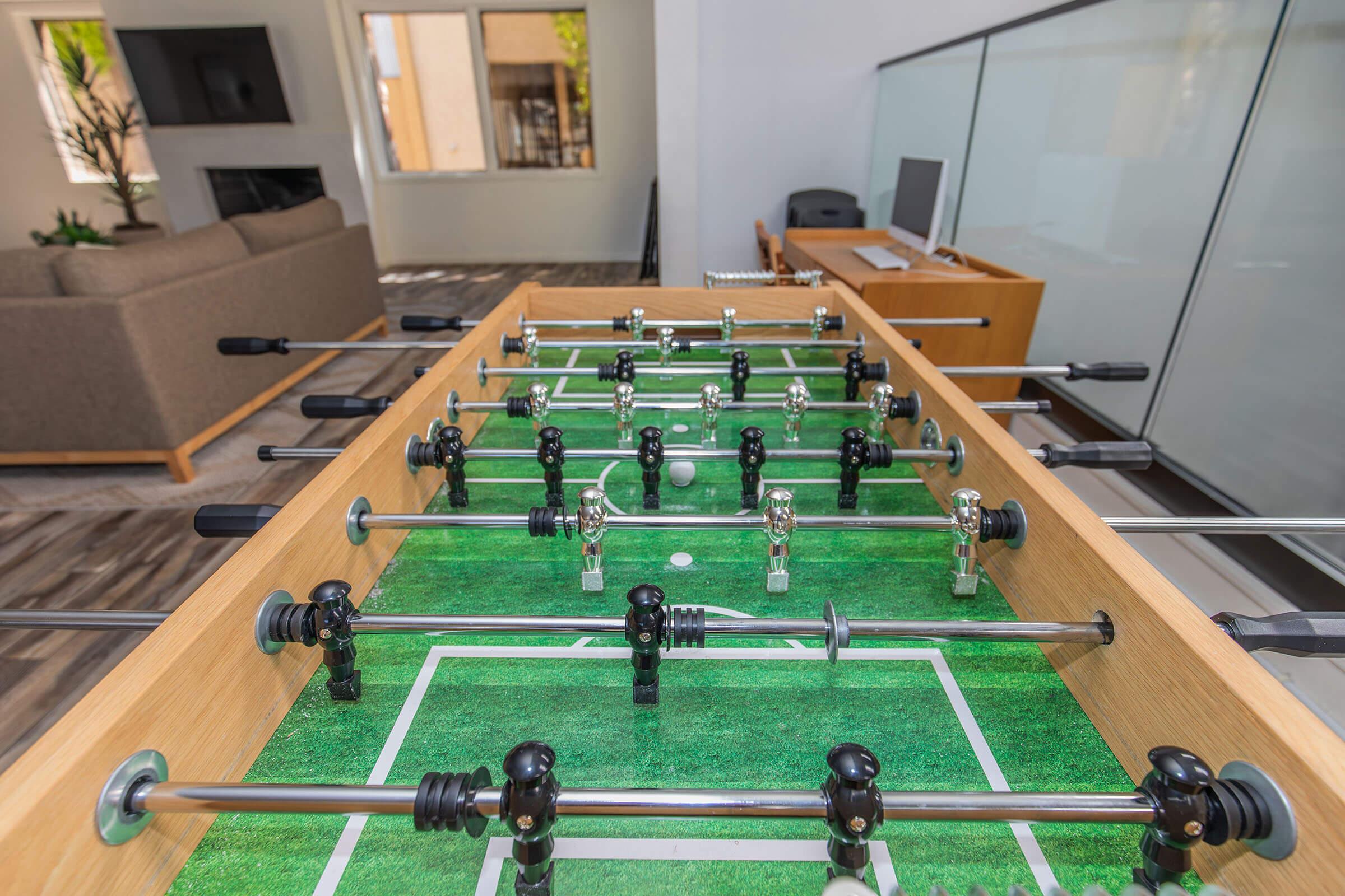 A foosball table with black and silver players positioned on a green felt surface, located in a bright room with a sofa, large windows, and a desk in the background. The setting is modern and inviting, perfect for a casual game with friends.