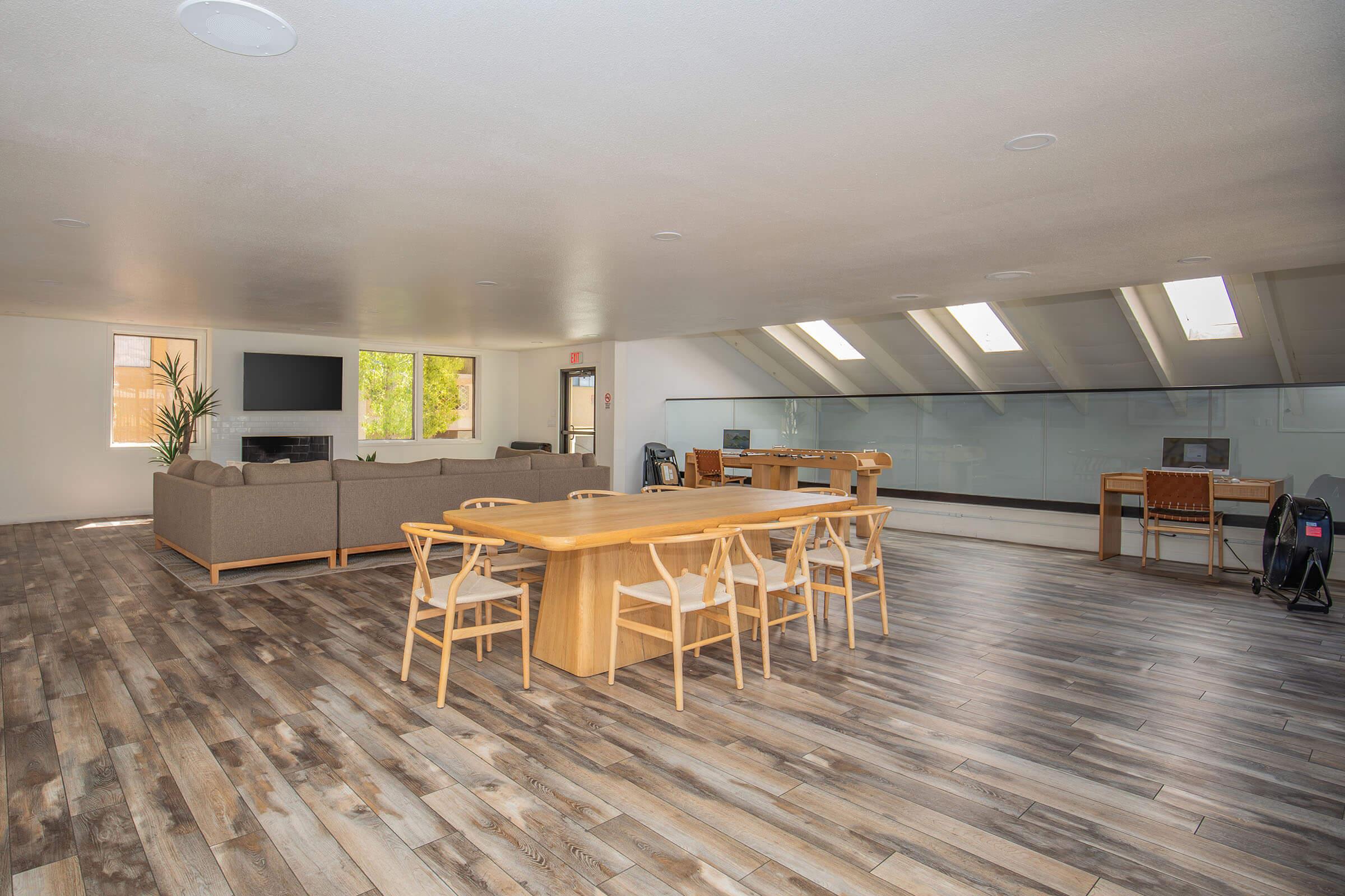 A spacious interior featuring a modern living area with a large grey sofa and a wooden dining table surrounded by white chairs. Large windows provide natural light, and there are two desk setups in the background. The floor is made of wooden planks, enhancing the contemporary design.