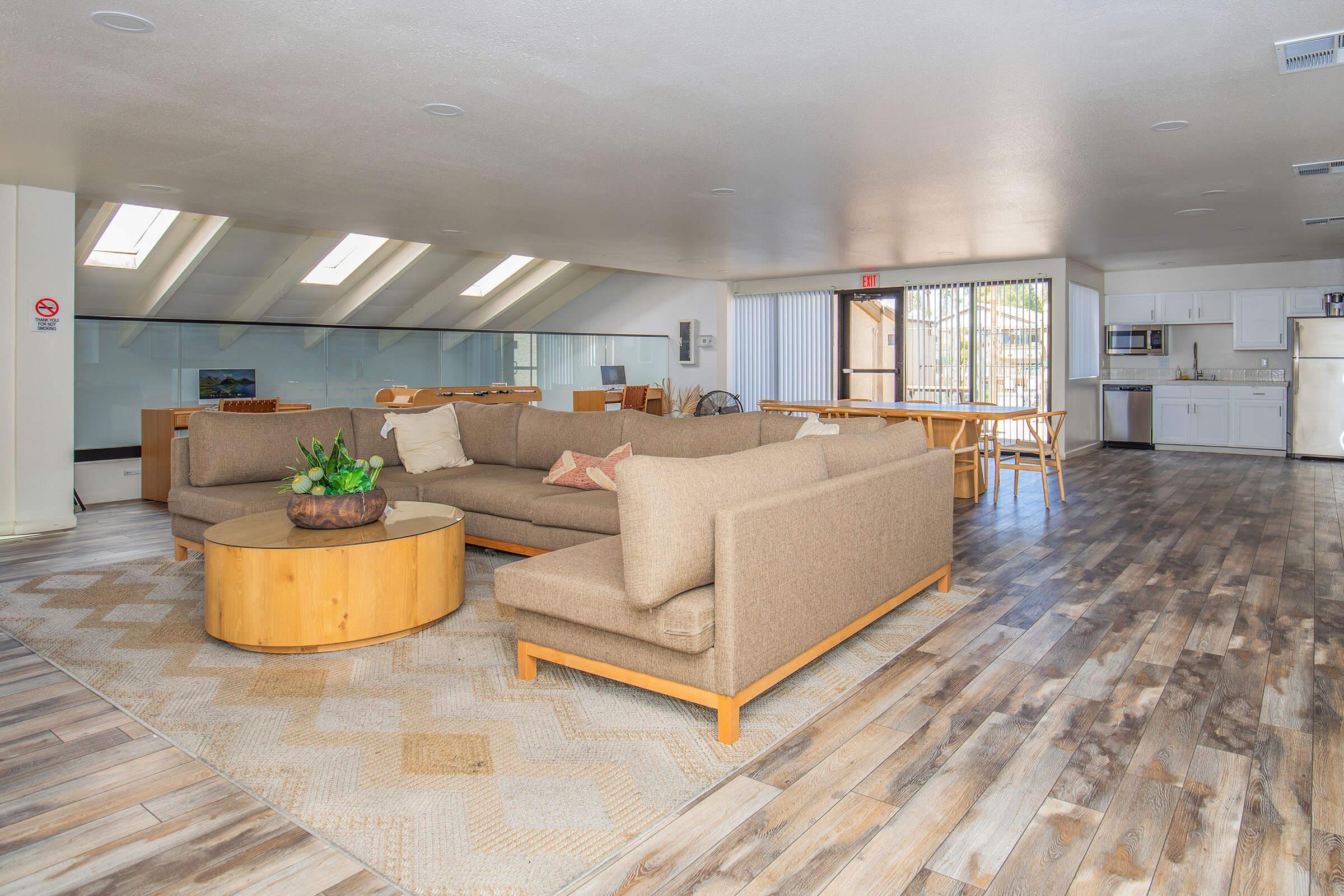 A spacious, modern living room featuring a large sectional sofa, a circular wooden coffee table with a plant centerpiece, and a light-colored area rug. The room has large windows allowing natural light, wooden flooring, and a dining area in the background with a kitchen visible.
