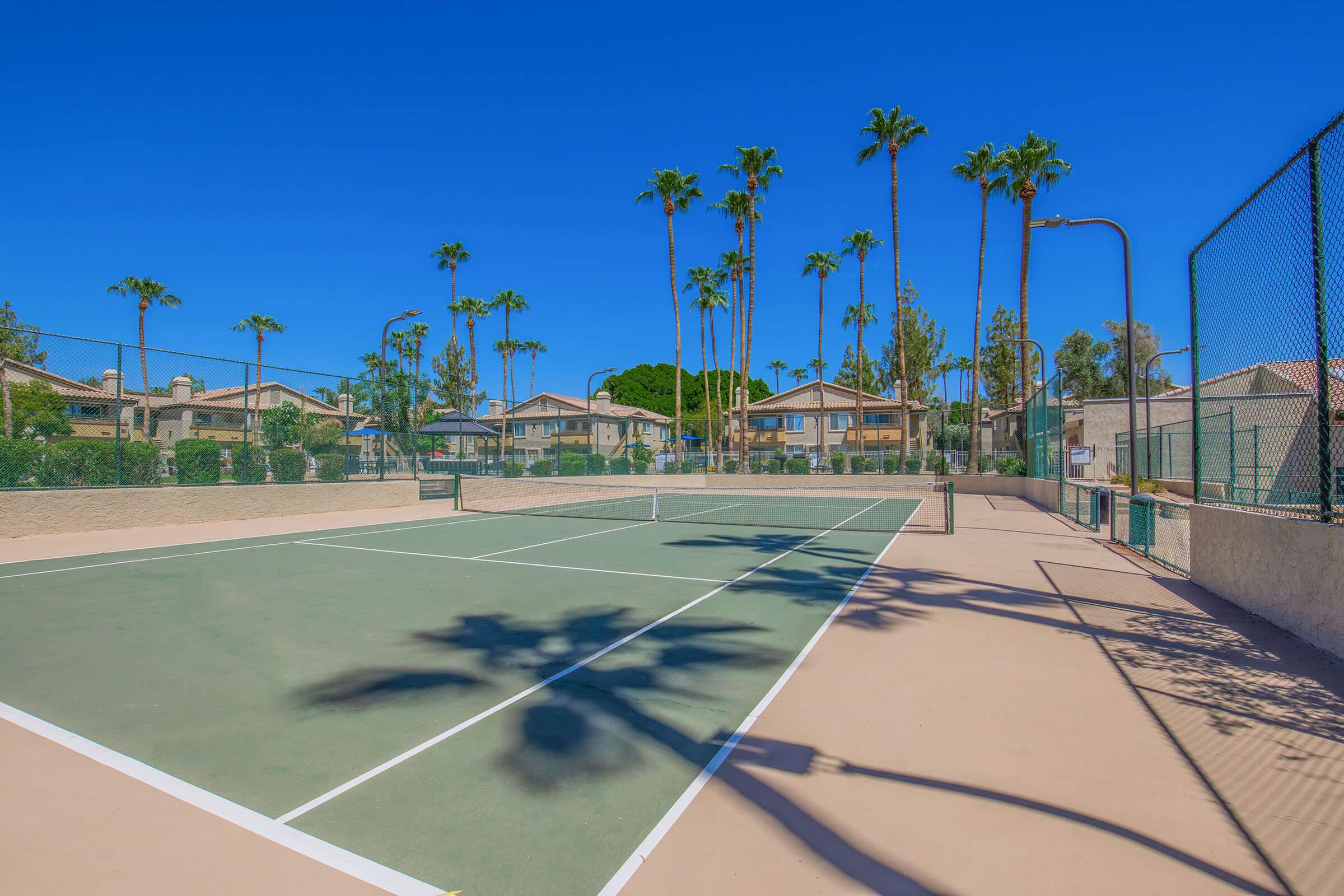 A well-maintained outdoor tennis court surrounded by palm trees and residential buildings. The blue sky provides a bright backdrop, while the court features a light-colored surface and clear green lines. The scene conveys a sunny, inviting atmosphere for sports and recreation.