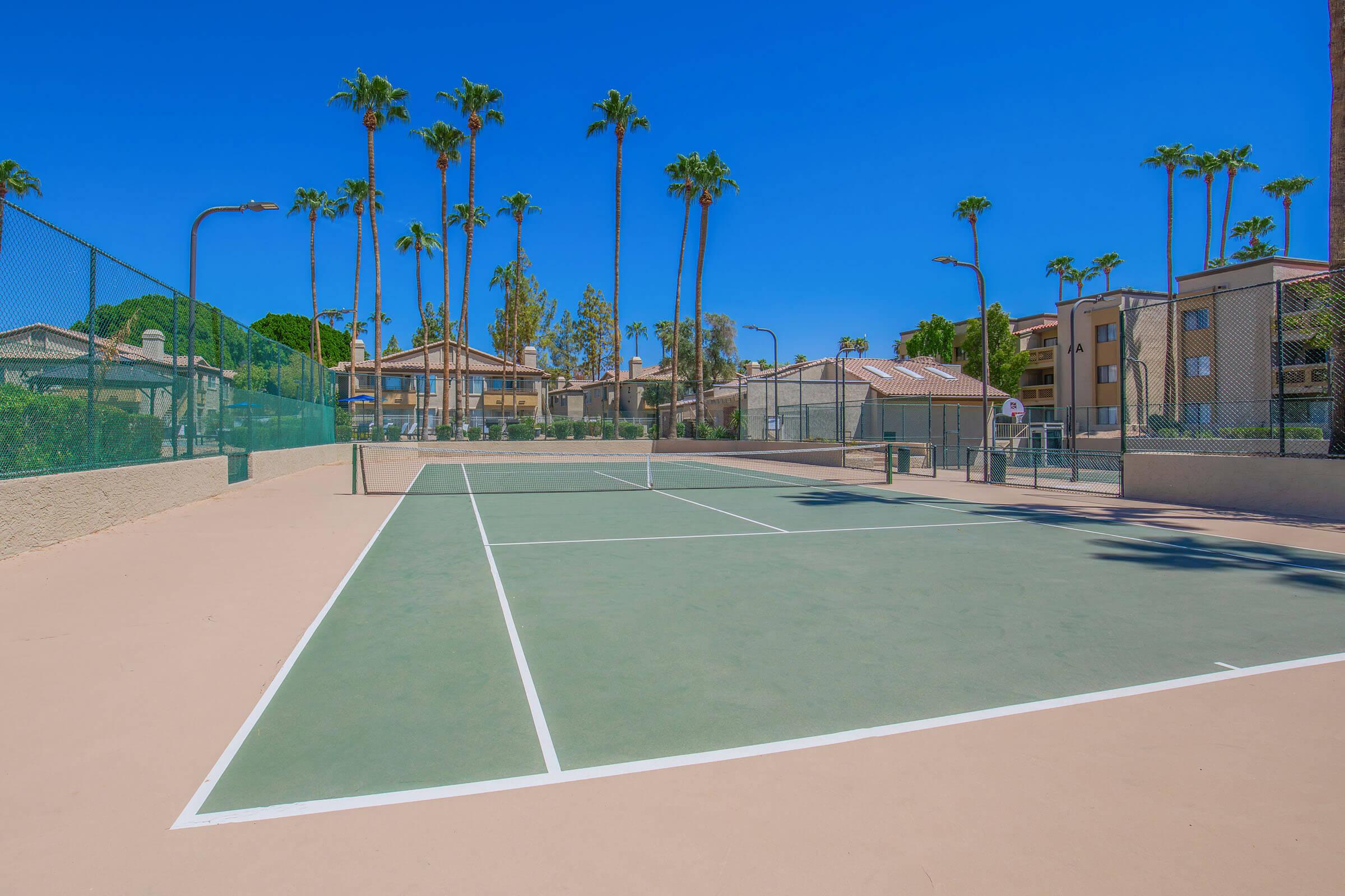 A well-maintained outdoor tennis court surrounded by tall palm trees under a clear blue sky. The court features a light-colored surface with white lines and is located near residential buildings in a sunny environment.