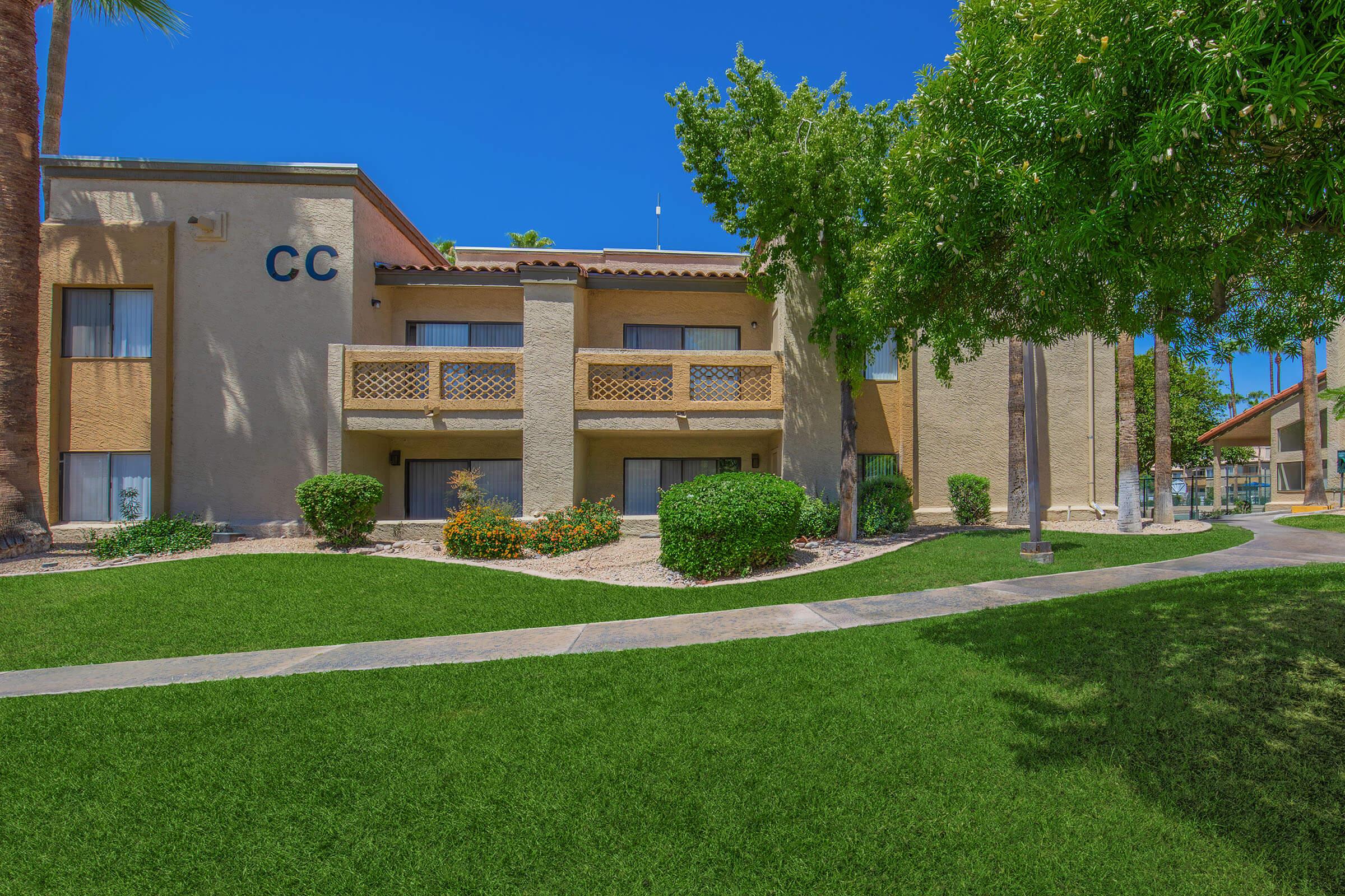 A two-story residential building with a beige exterior featuring a balcony, surrounded by green grass and landscaped shrubs. Palm trees frame the area under a clear blue sky.
