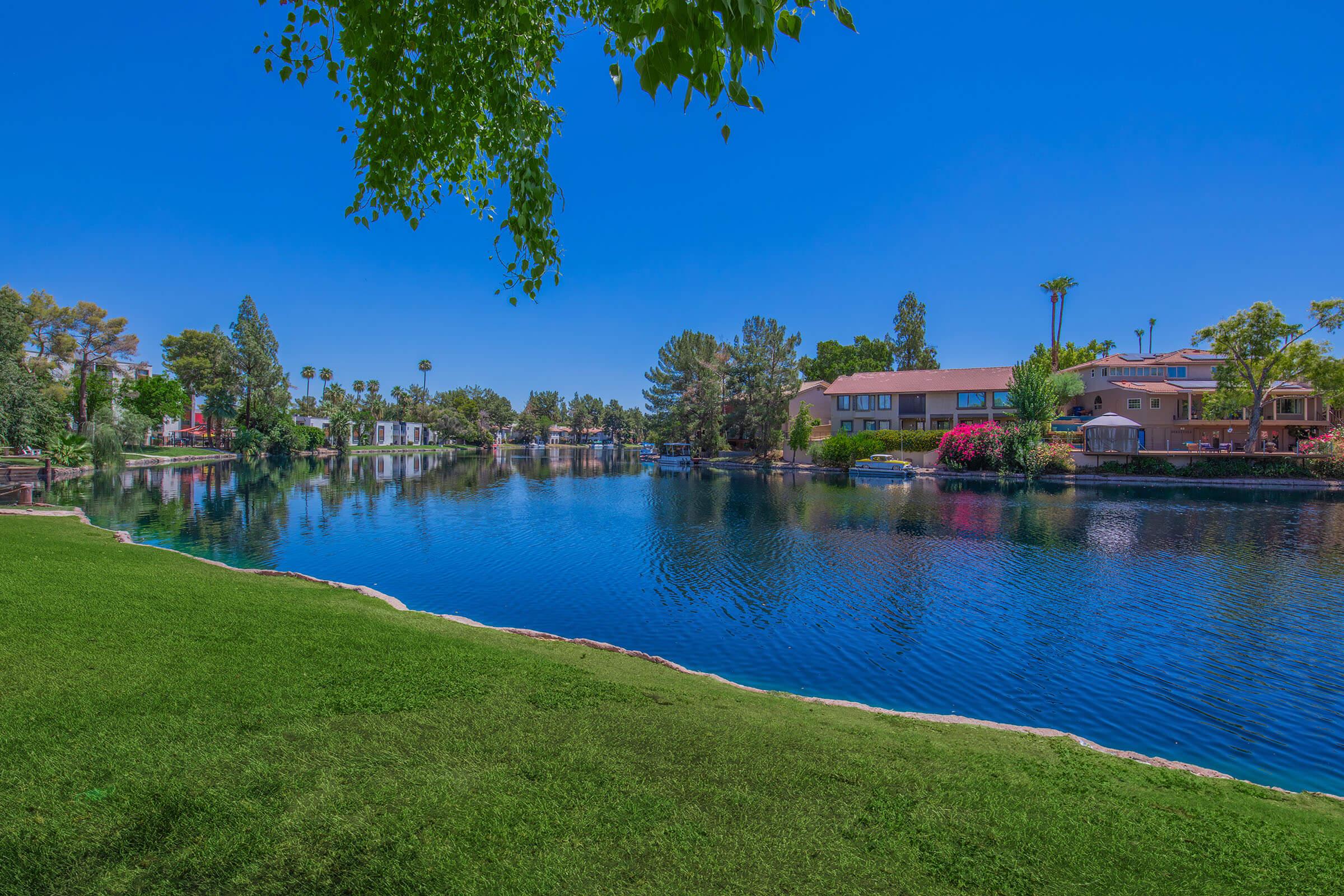 A serene lake surrounded by lush green grass and trees, with colorful houses lining the shore. The clear blue sky reflects on the water, creating a picturesque and tranquil scene. Bright flowers add a pop of color to the landscape, enhancing the peaceful atmosphere.