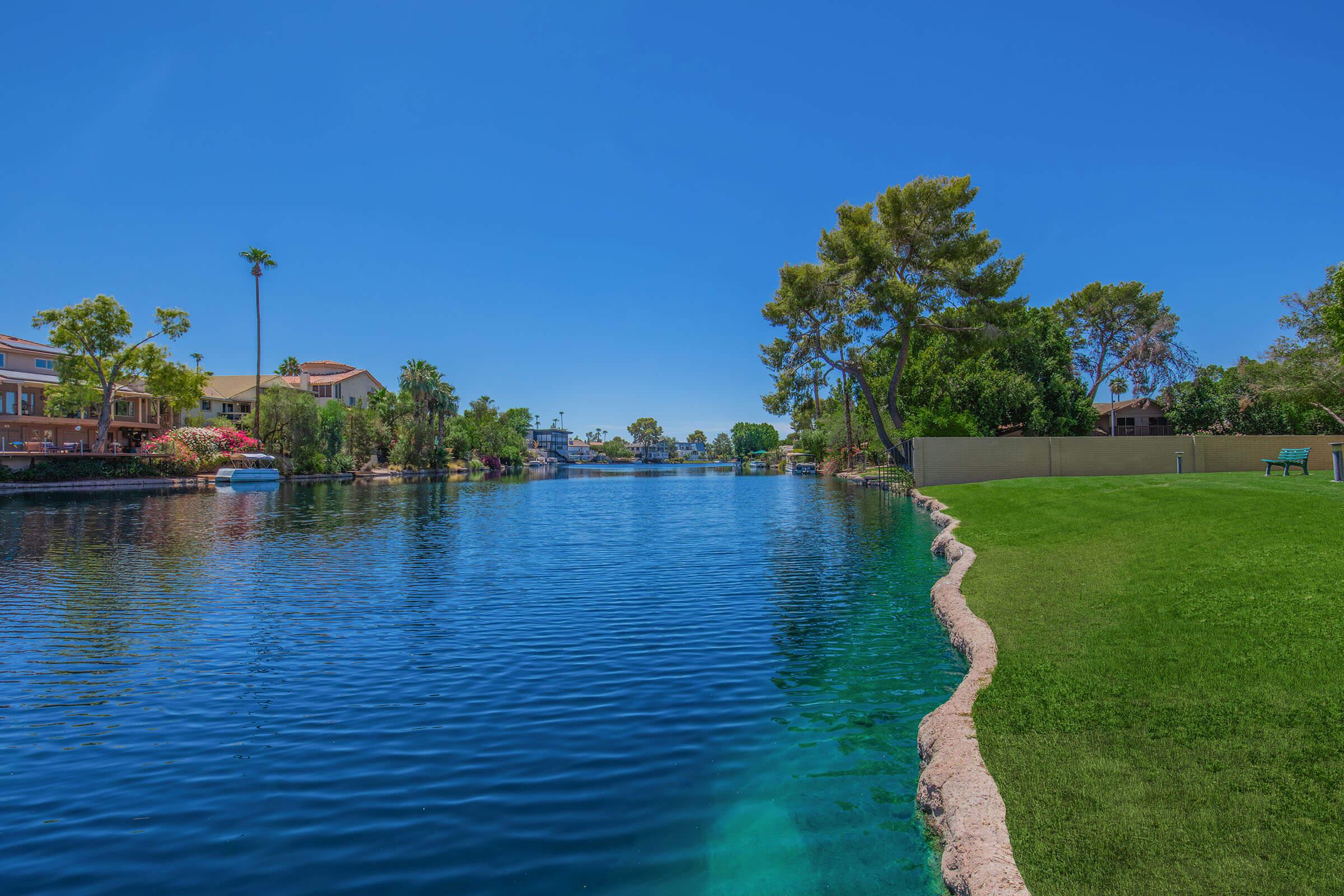 A serene lake scene features clear blue water bordered by lush green grass and trees. On one side, there are residential homes with colorful landscaping, while the opposite side showcases more greenery. The bright sky and calm waters create a tranquil atmosphere, ideal for relaxation and recreation.