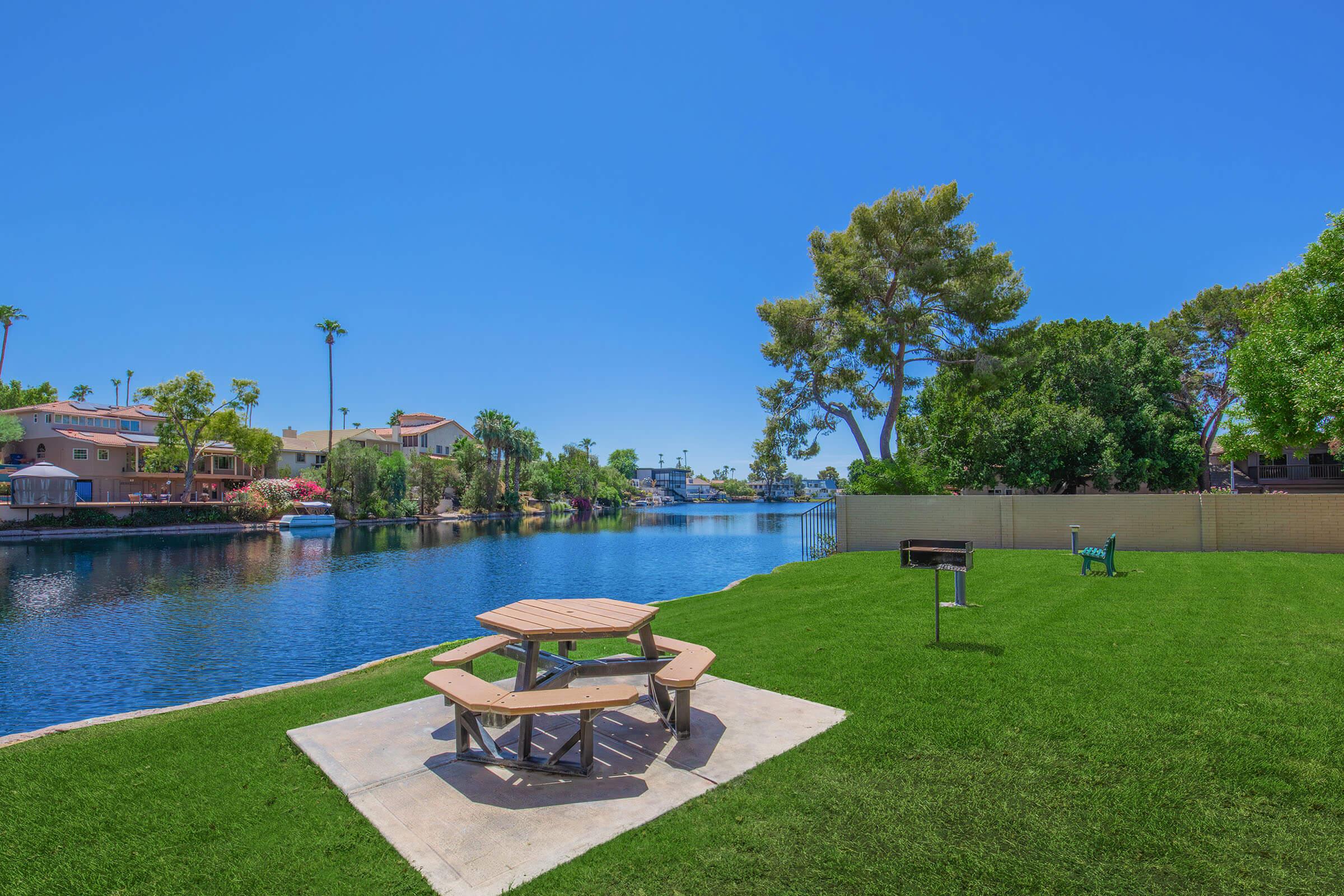 A serene lakeside scene featuring a picnic table on a green lawn, with a barbecue grill nearby. The calm water reflects the clear blue sky, surrounded by lush trees and residential houses in the background, creating a peaceful outdoor space.