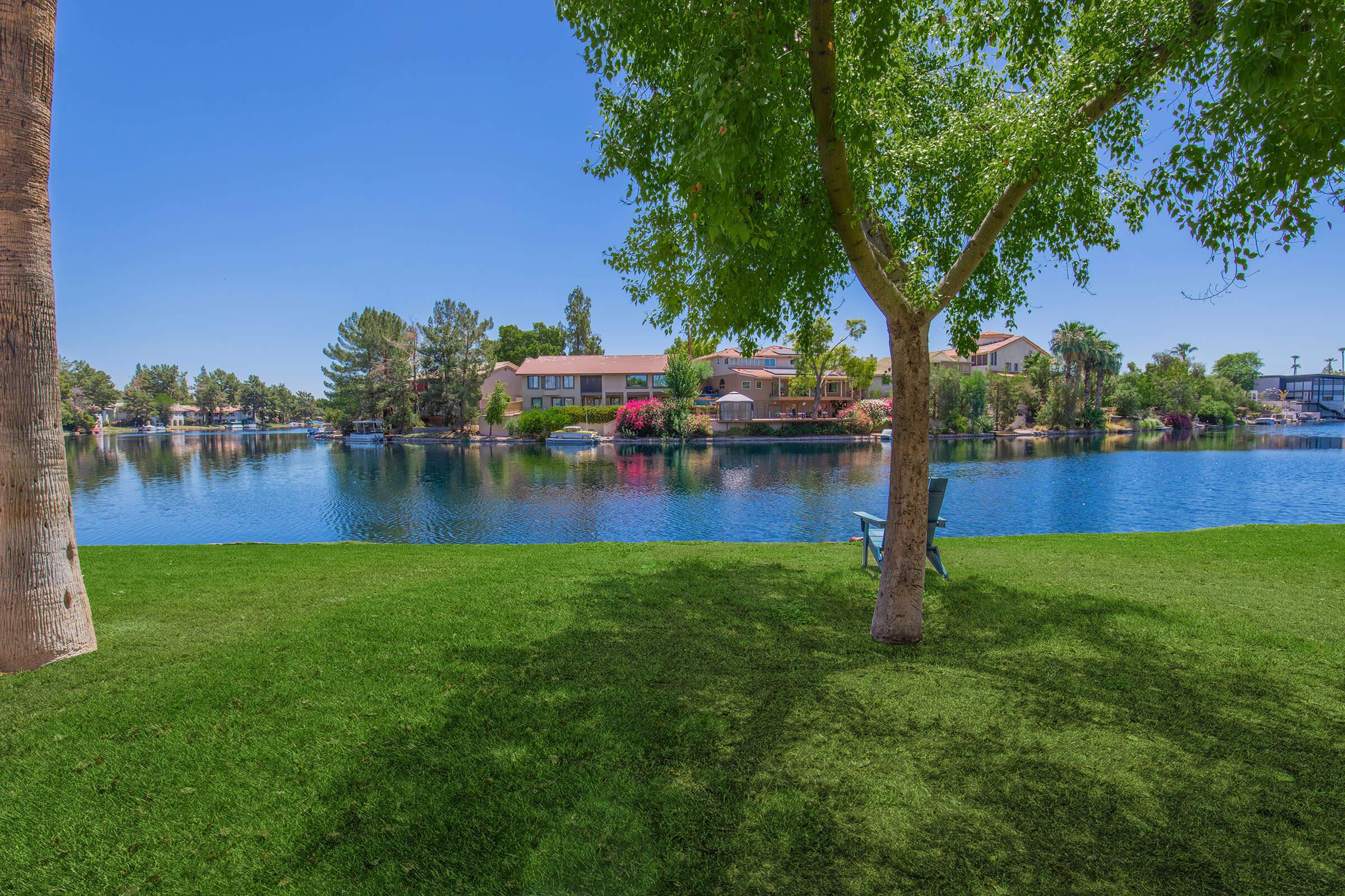 A picturesque lakeside scene featuring vibrant green grass, a large tree providing shade, and a calm blue lake reflecting nearby houses and greenery under a clear blue sky. A single lawn chair is positioned on the grass, inviting relaxation and enjoyment of the serene landscape.