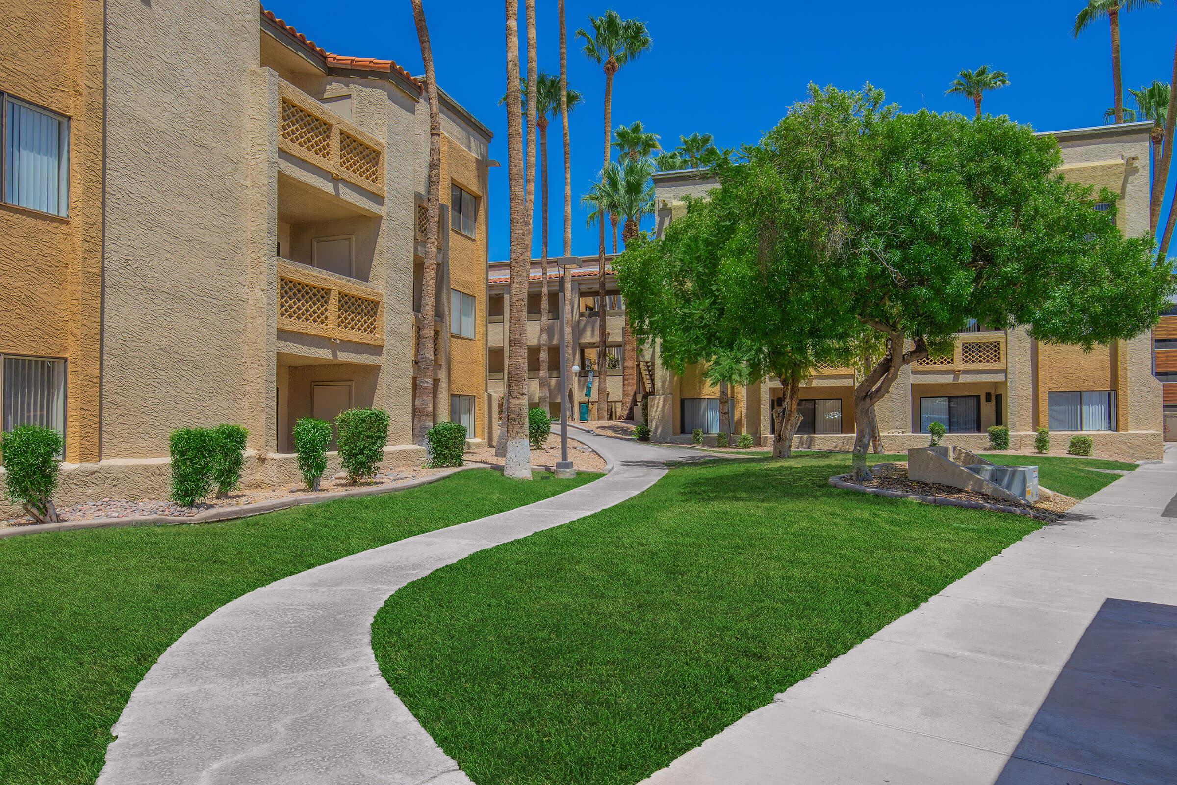 A sunny courtyard scene featuring two multi-story apartment buildings painted in beige, surrounded by well-kept landscaping. A winding pathway leads through lush green lawns and palm trees, creating a welcoming outdoor space. Clear blue sky enhances the vibrant atmosphere.