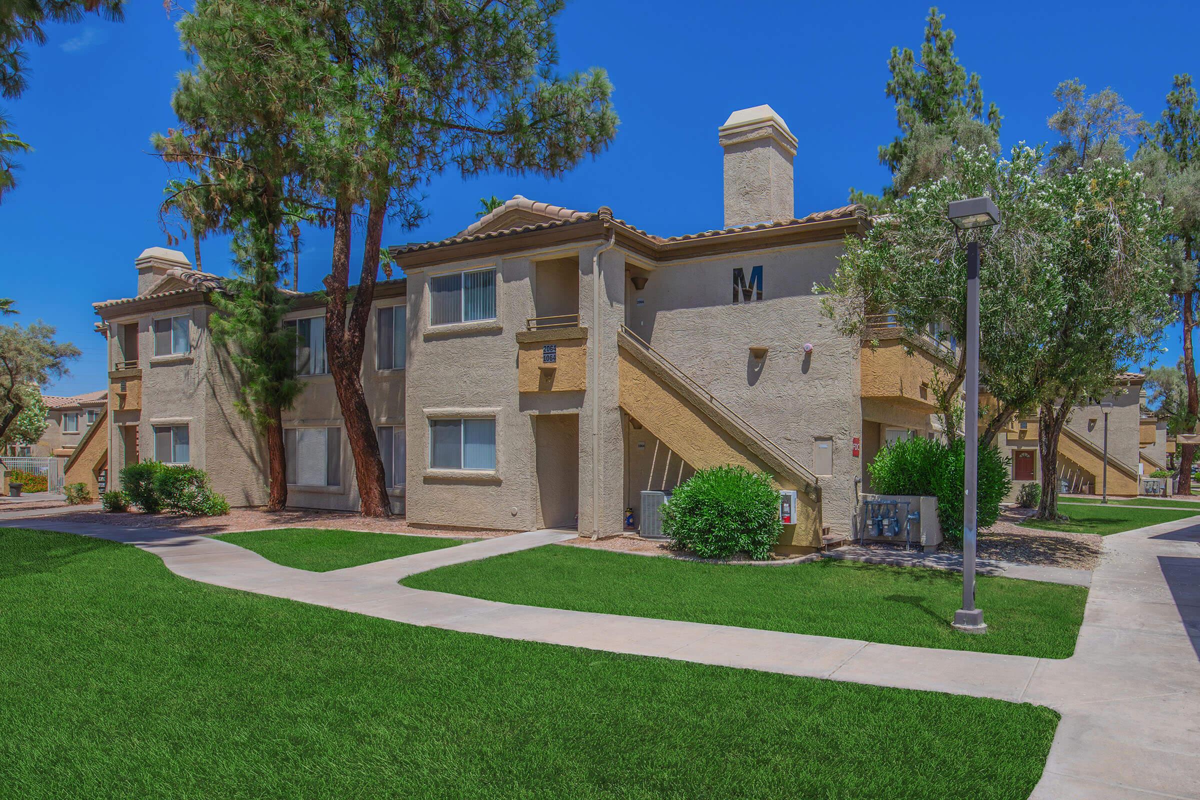 A bright, sunny view of a residential apartment complex featuring two-story buildings with light-colored exteriors. The well-maintained grounds include lush green grass, neatly trimmed bushes, and trees, with a clear blue sky overhead. Walking paths are visible in front of the buildings.
