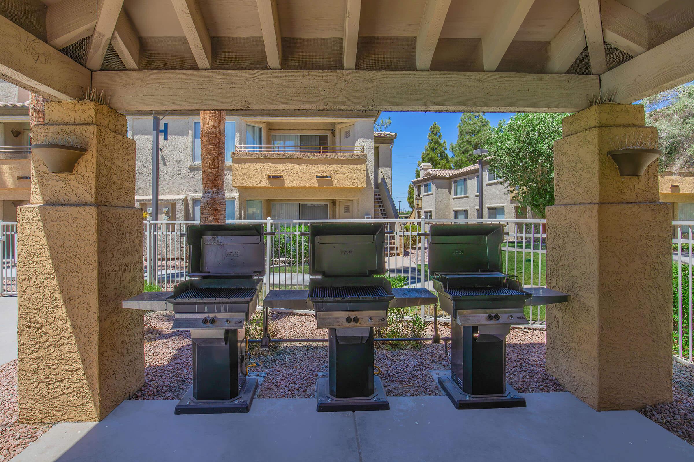 Outdoor grilling area featuring three black gas grills under a covered patio. The setting includes stone columns, a concrete path, and a fenced lawn area with residential buildings in the background. Bright, sunny day with trees and shrubs nearby.