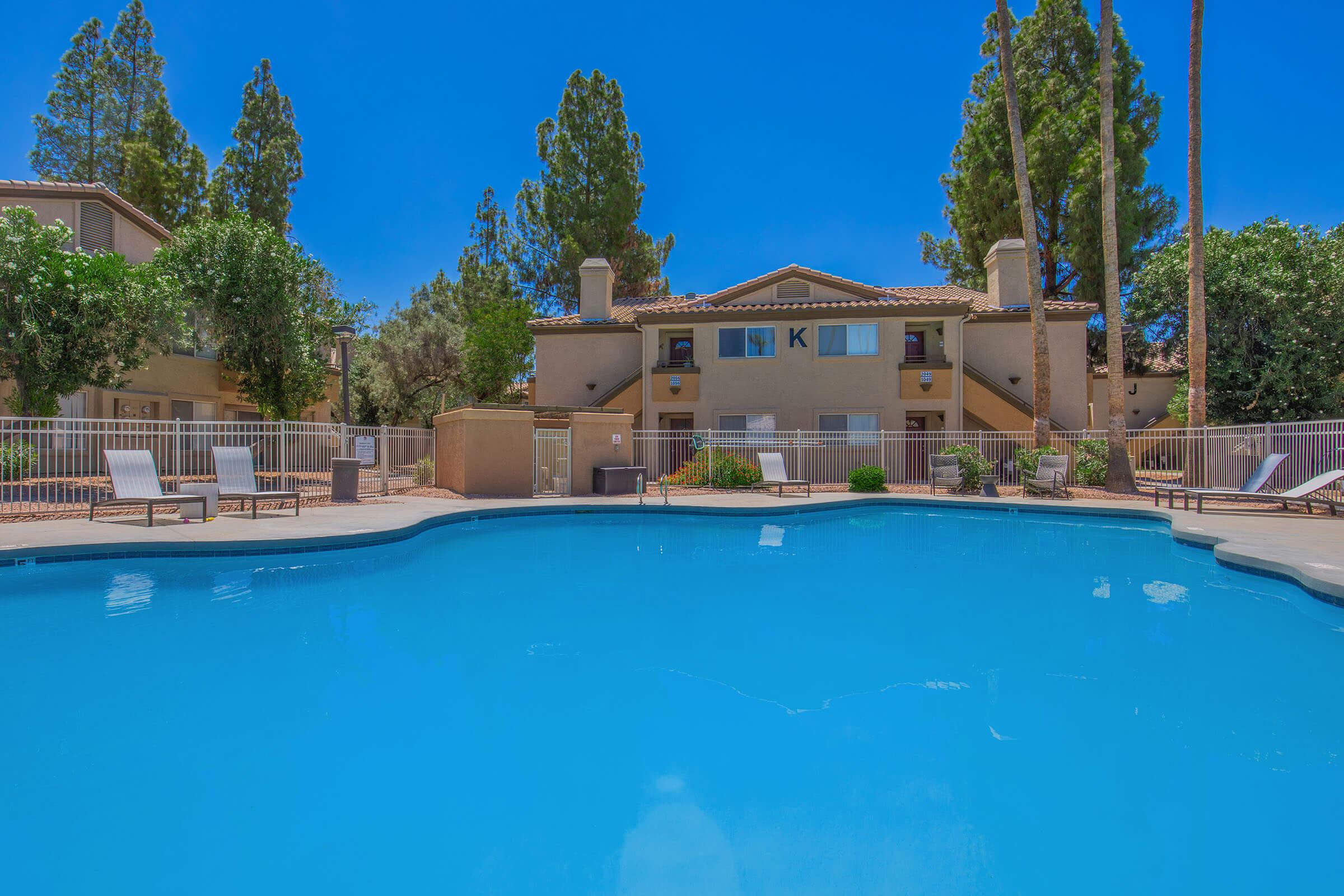 A serene swimming pool surrounded by lounge chairs, with lush greenery and tall trees in the background. In the distance, a two-story building with balconies is visible under a clear blue sky. The scene conveys a tranquil, inviting atmosphere perfect for relaxation.