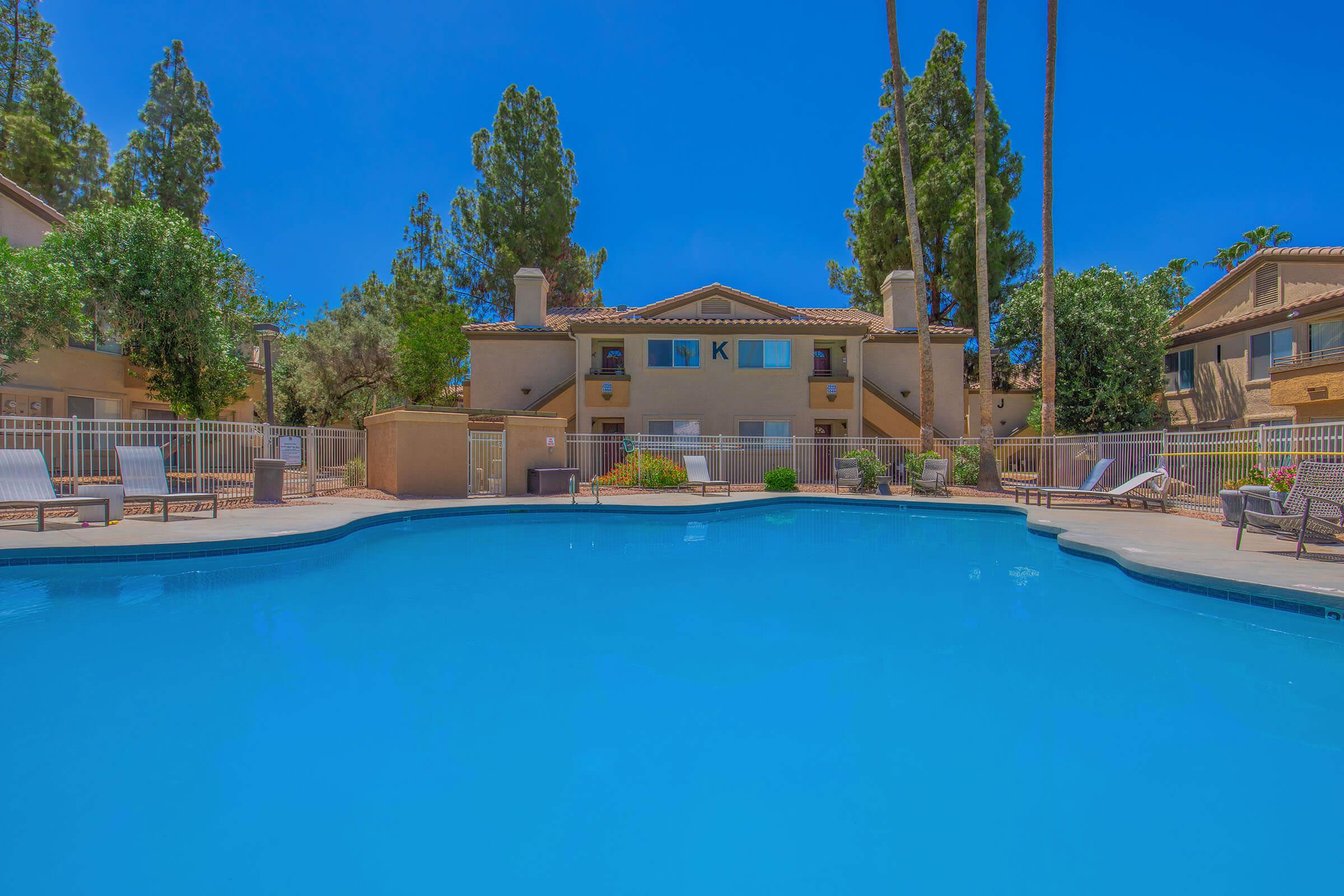 A swimming pool surrounded by lounge chairs, with a backdrop of landscaped trees and residential buildings. The sky is clear and blue, creating a serene outdoor atmosphere for relaxation and leisure.