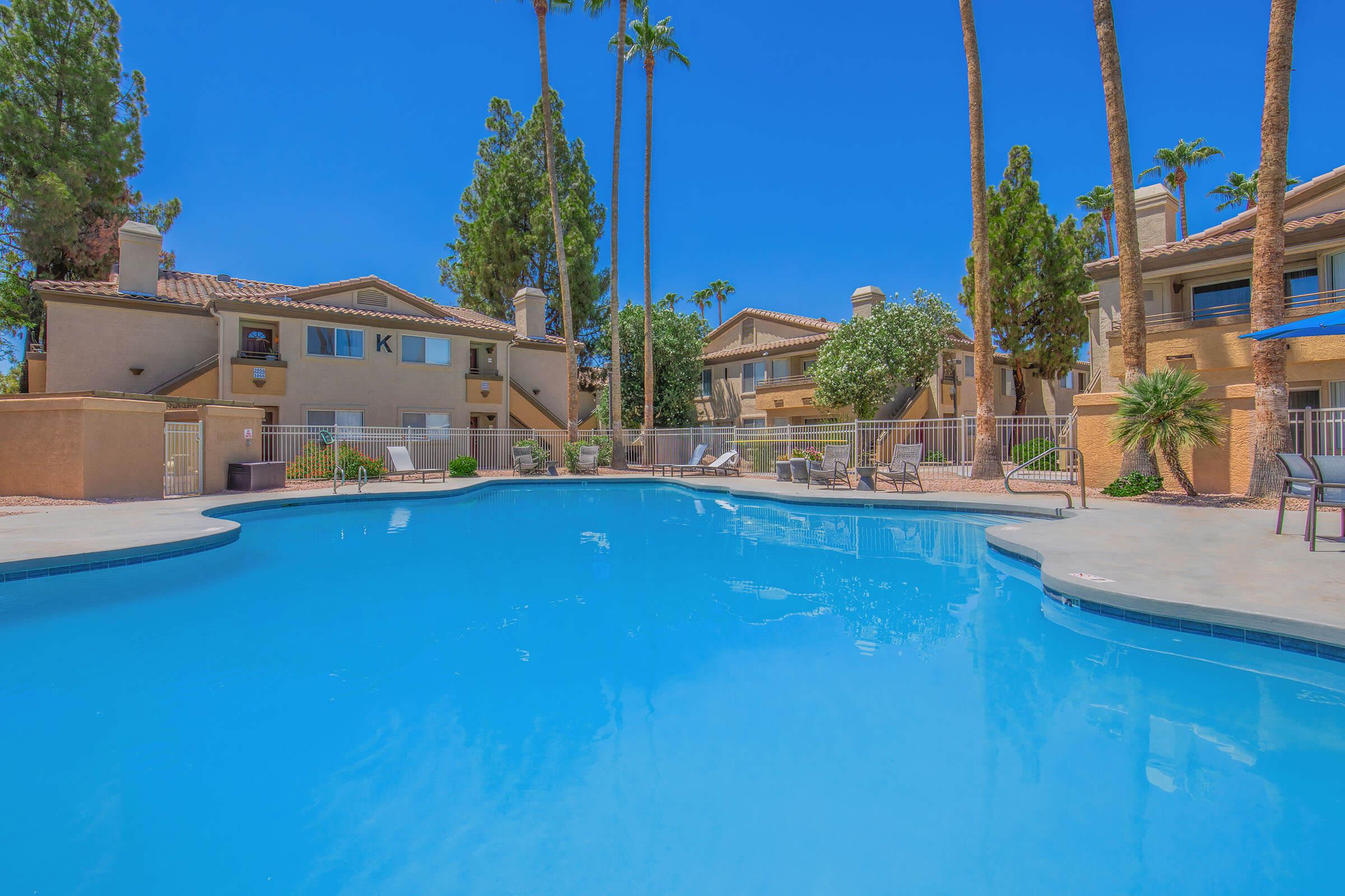 A bright blue swimming pool surrounded by tall palm trees and landscaped grounds. In the background, there are beige apartment buildings with balconies and patio furniture. The sky is clear and blue, creating a vibrant and inviting atmosphere.
