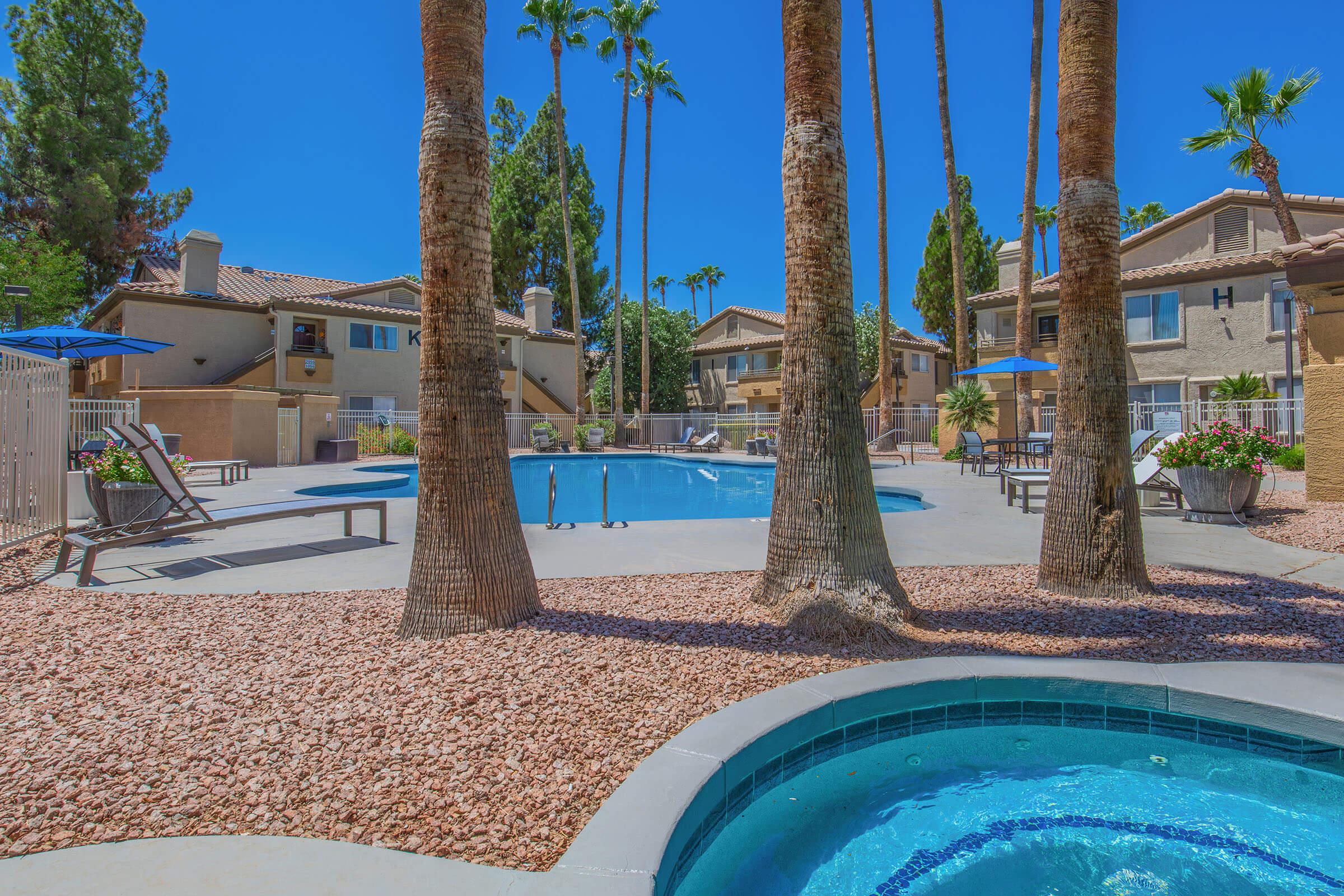A bright, sunny pool area surrounded by palm trees and landscaped with gravel. The pool features lounge chairs and umbrellas, with residential buildings in the background. A hot tub is visible in the foreground, enhancing the relaxing atmosphere of the outdoor space.