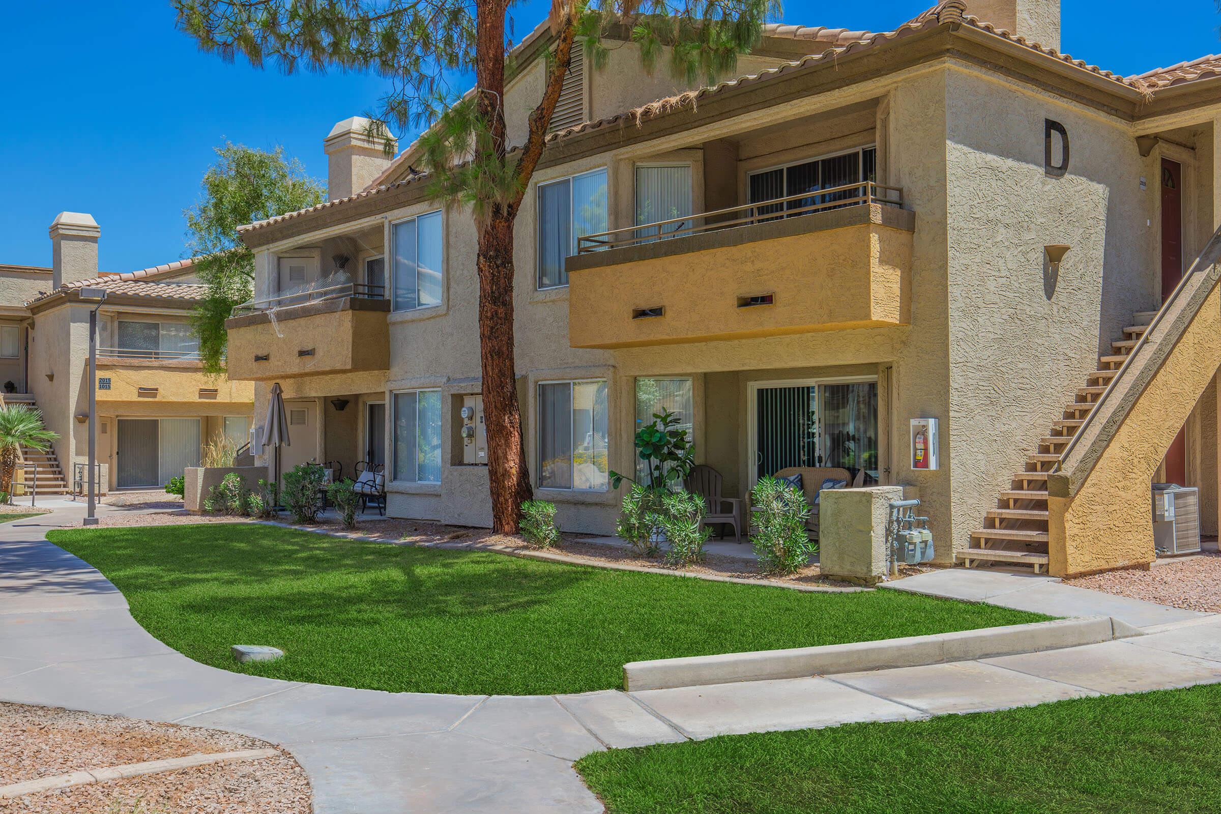 Two-story residential building featuring multiple apartments, with a light beige facade and balconies. A green lawn and concrete pathway are in the foreground, flanked by palm trees and decorative shrubs. Clear blue sky above enhances the bright, sunny atmosphere.